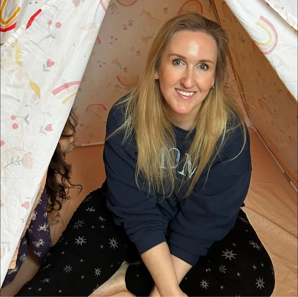Young woman with long blonde hair smiling, sitting inside a colorful fabric tent with floral and rainbow patterns, with another person partially visible behind her.
