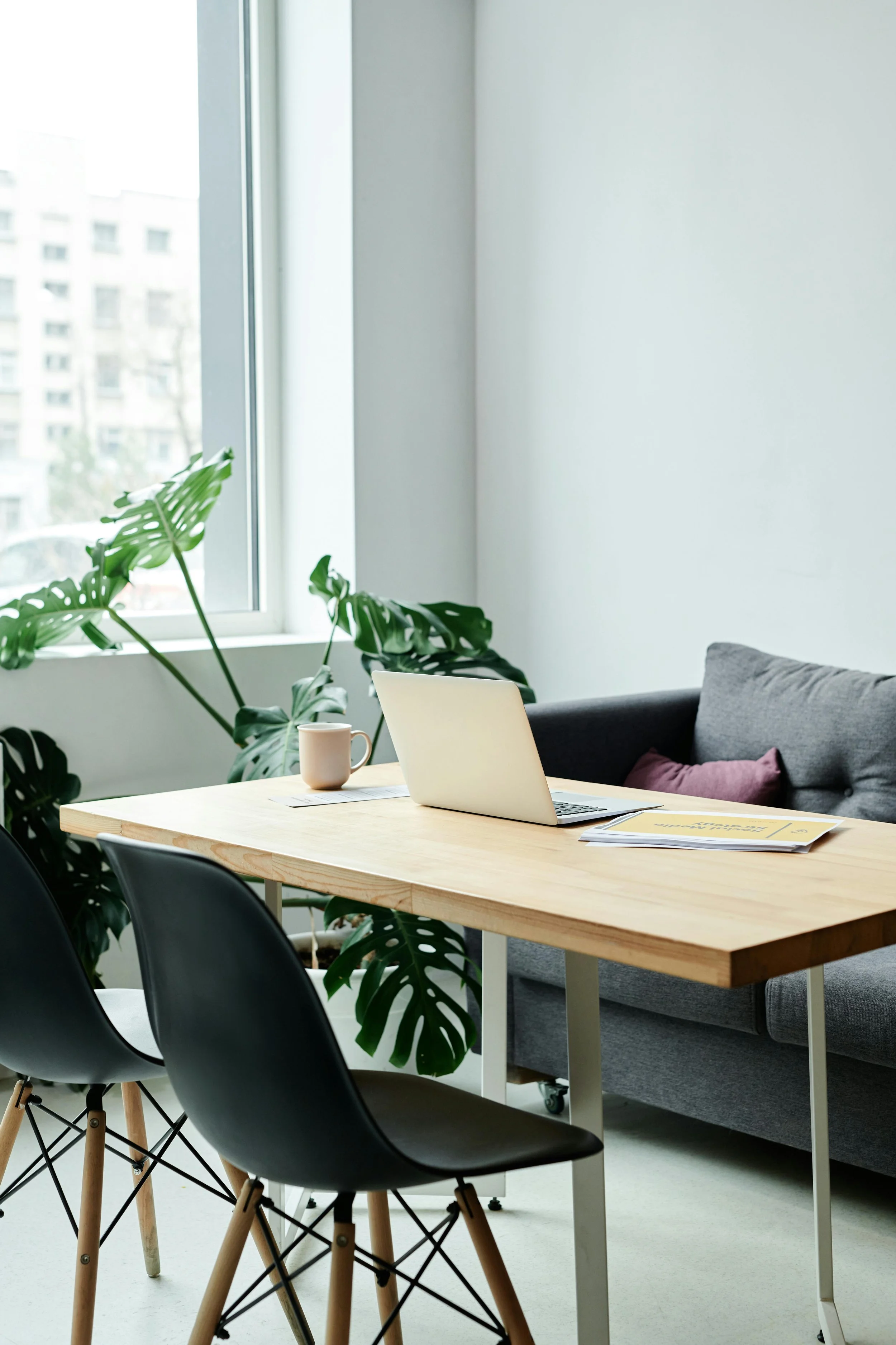 A bright modern office space with a wooden table, black chairs, a laptop, a beige mug, and papers. In the background, there is a sofa with pillows, large windows with a view of buildings, and some green houseplants.