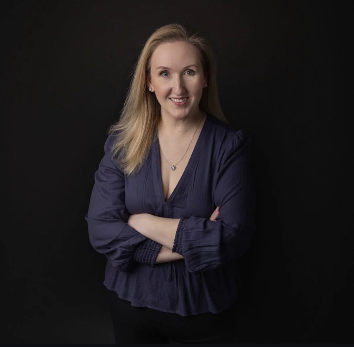 Portrait of a smiling woman with blonde hair, wearing a navy blue blouse with a V-neck and a necklace, standing against a black background.
