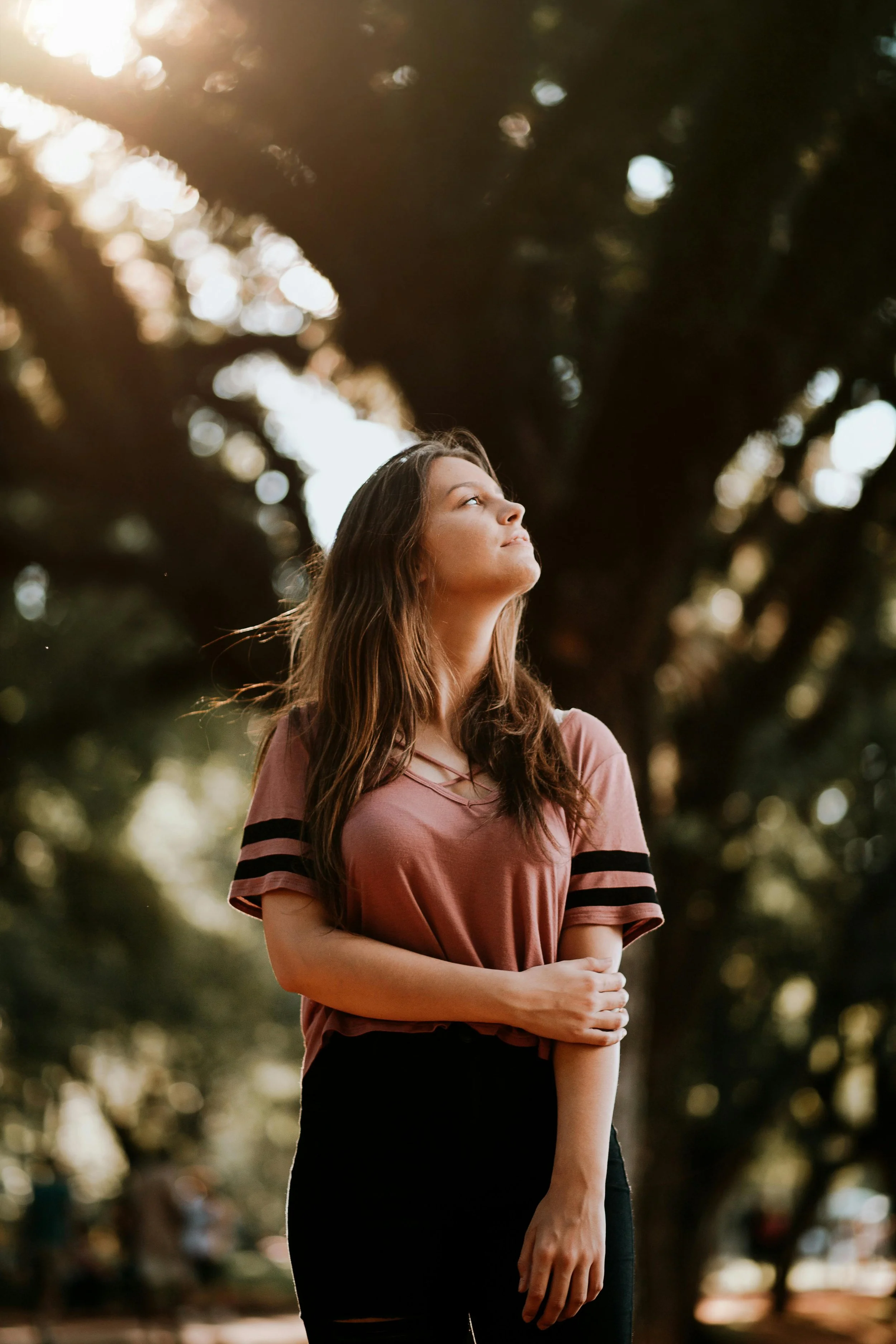 Young woman with long brown hair stands outdoors in sunlight, looking upwards with arms crossed, surrounded by trees.