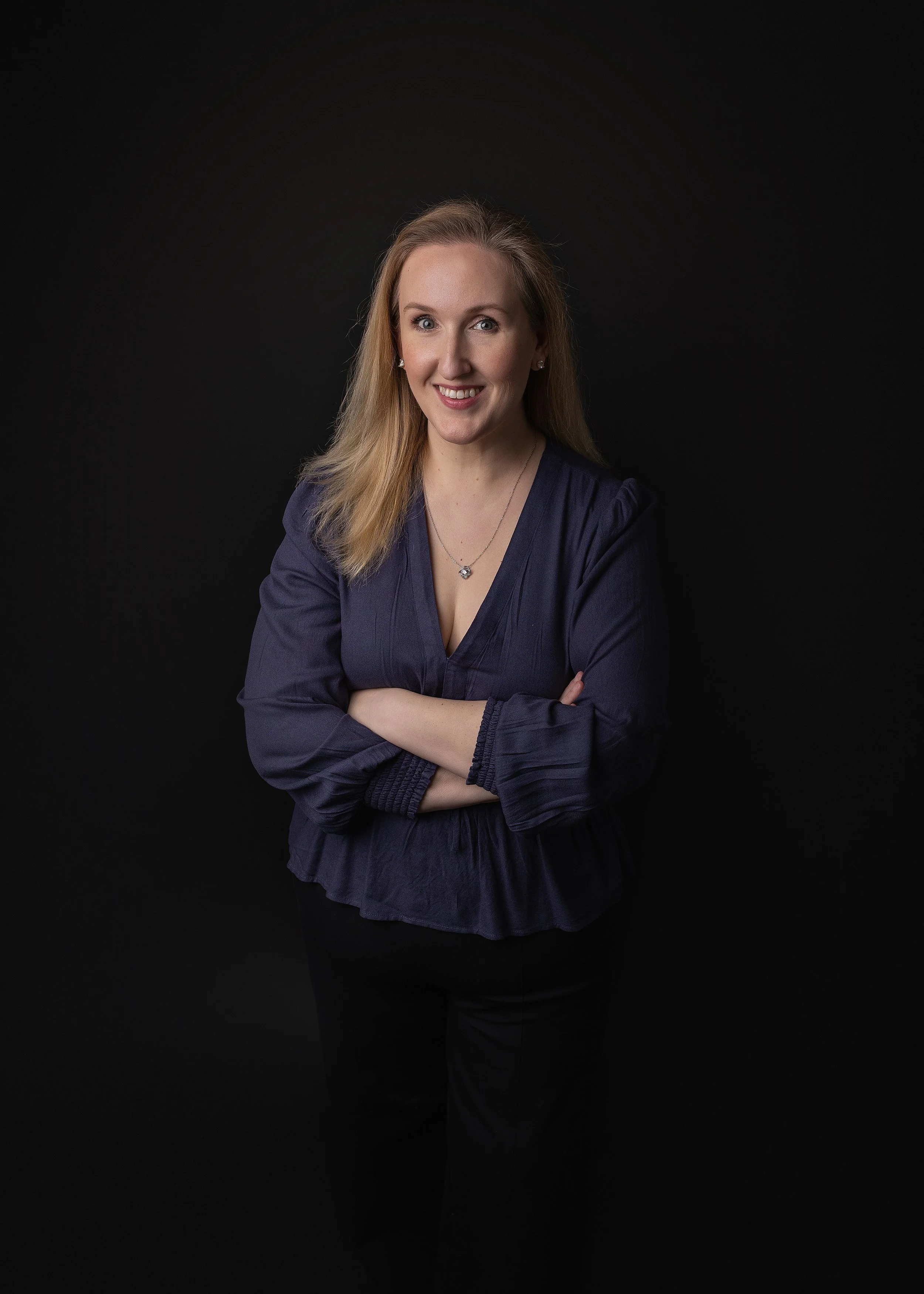 A woman with blonde hair wearing a navy blue blouse, posing against a black background with arms crossed and smiling at the camera.