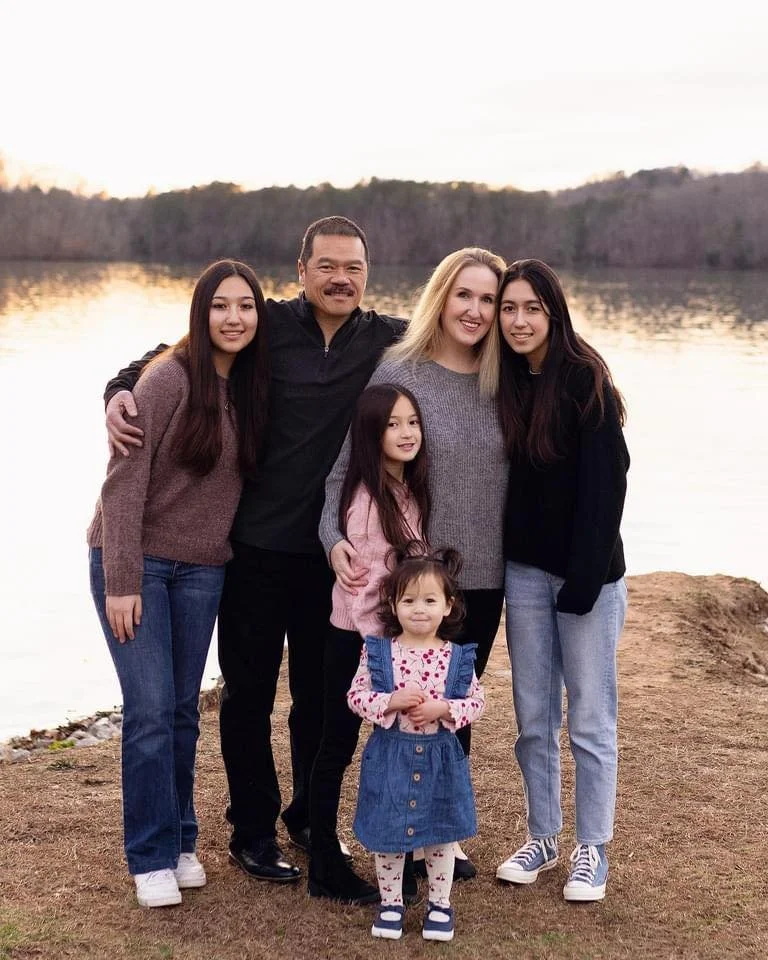 Family of six standing near a lake during sunset, smiling at the camera. The group includes two adults, three girls, and a young girl with curly hair, all dressed casually.