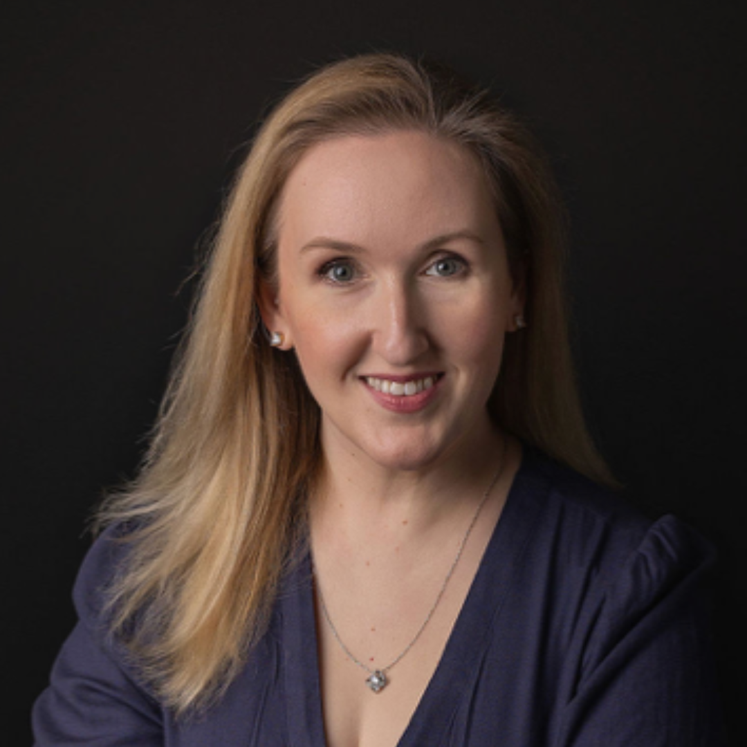 A woman with blonde hair and blue eyes smiling against a black background, wearing a dark blue top and silver jewelry.