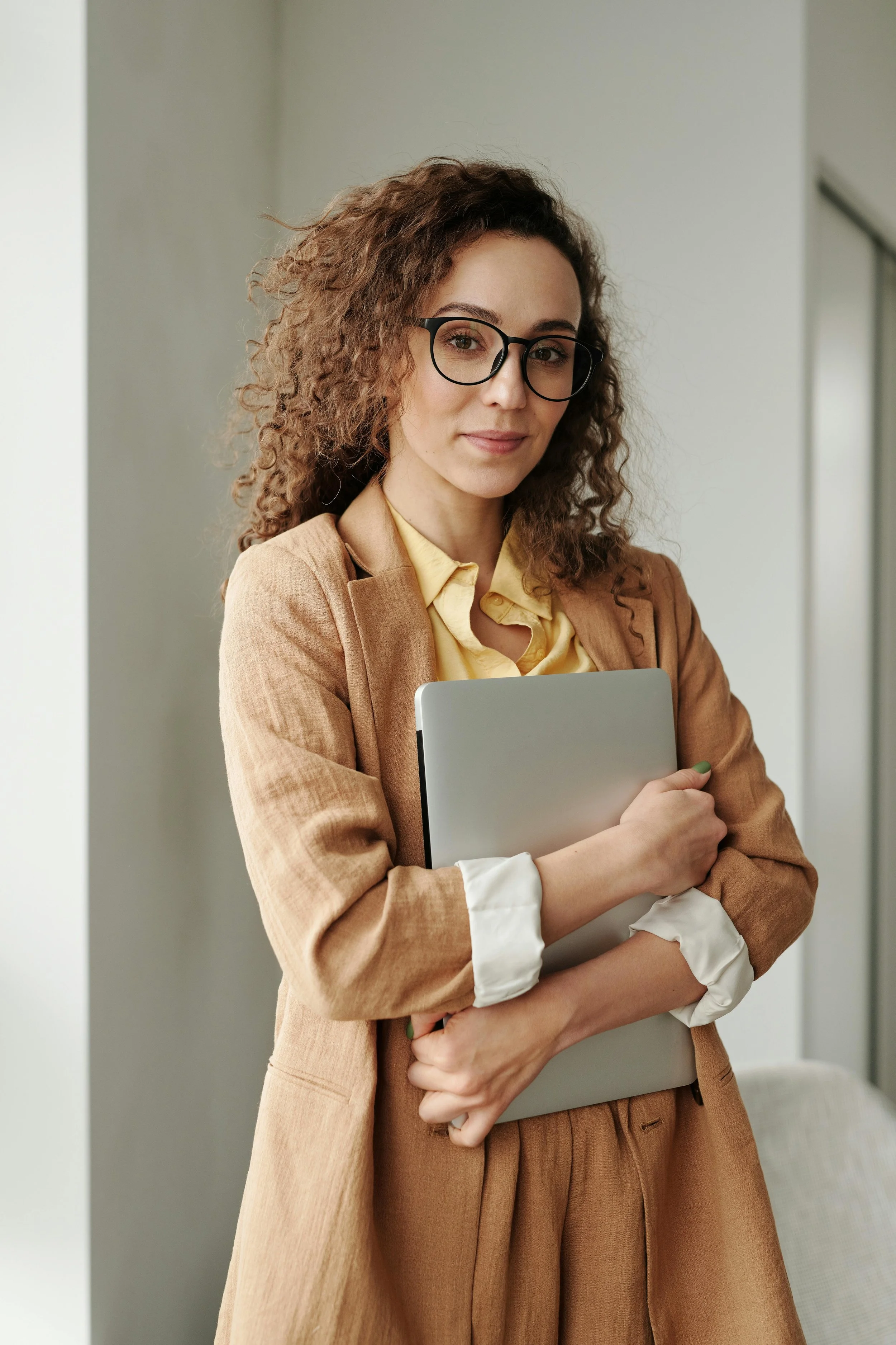 A woman with curly brown hair and black glasses, wearing a tan blazer over a yellow shirt, holding a laptop close to her chest with a slight smile, standing in a neutral background.