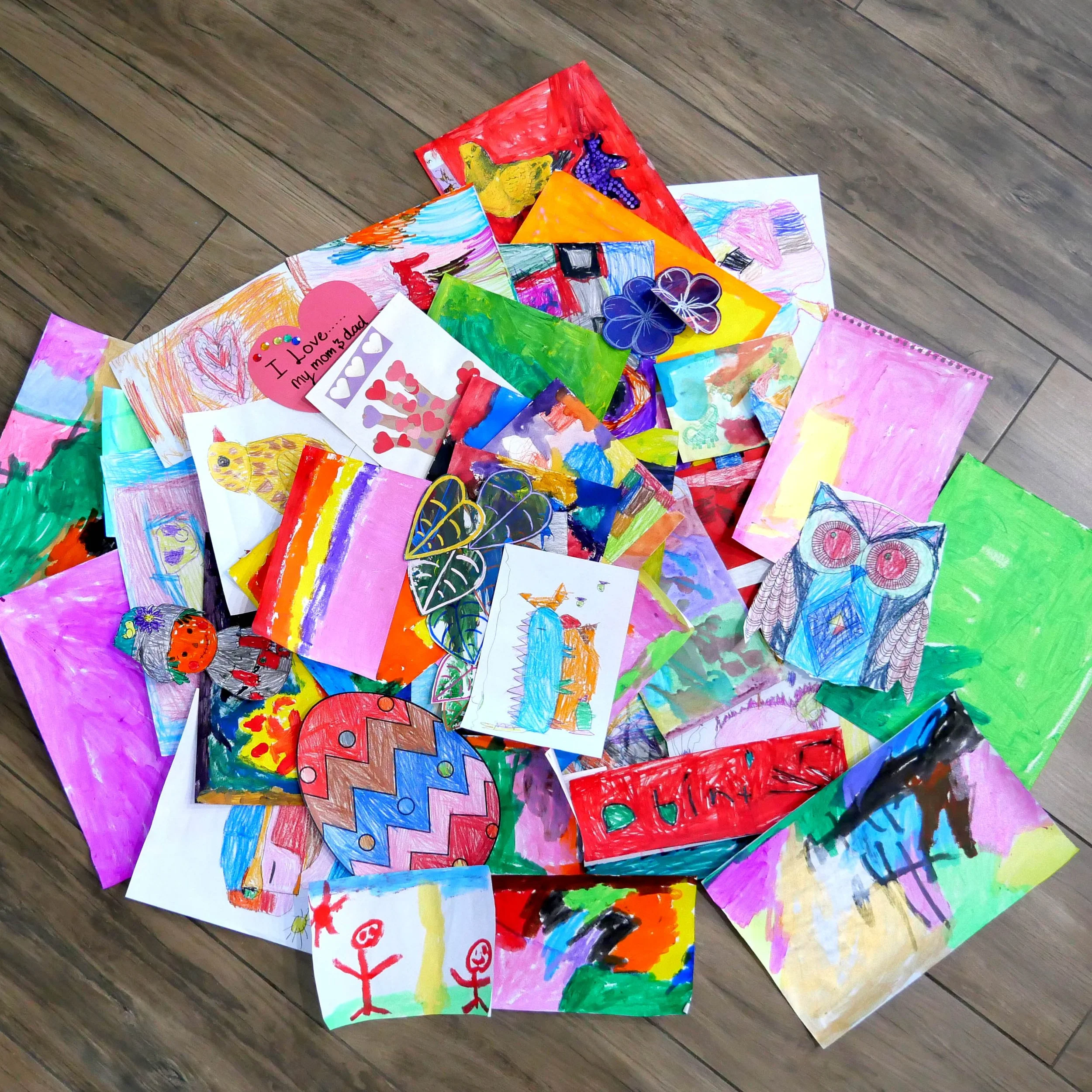 A collection of colorful children's drawings and paintings spread out on a wooden floor. The artwork includes various vibrant images such as hearts, animals, flowers, and abstract designs.