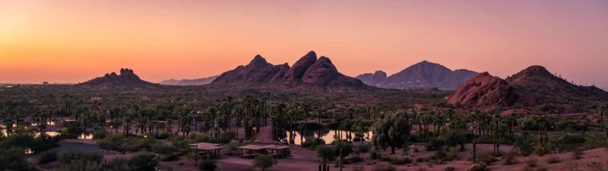 Sunset over rocky desert mountains with lakes and palm trees in the foreground.