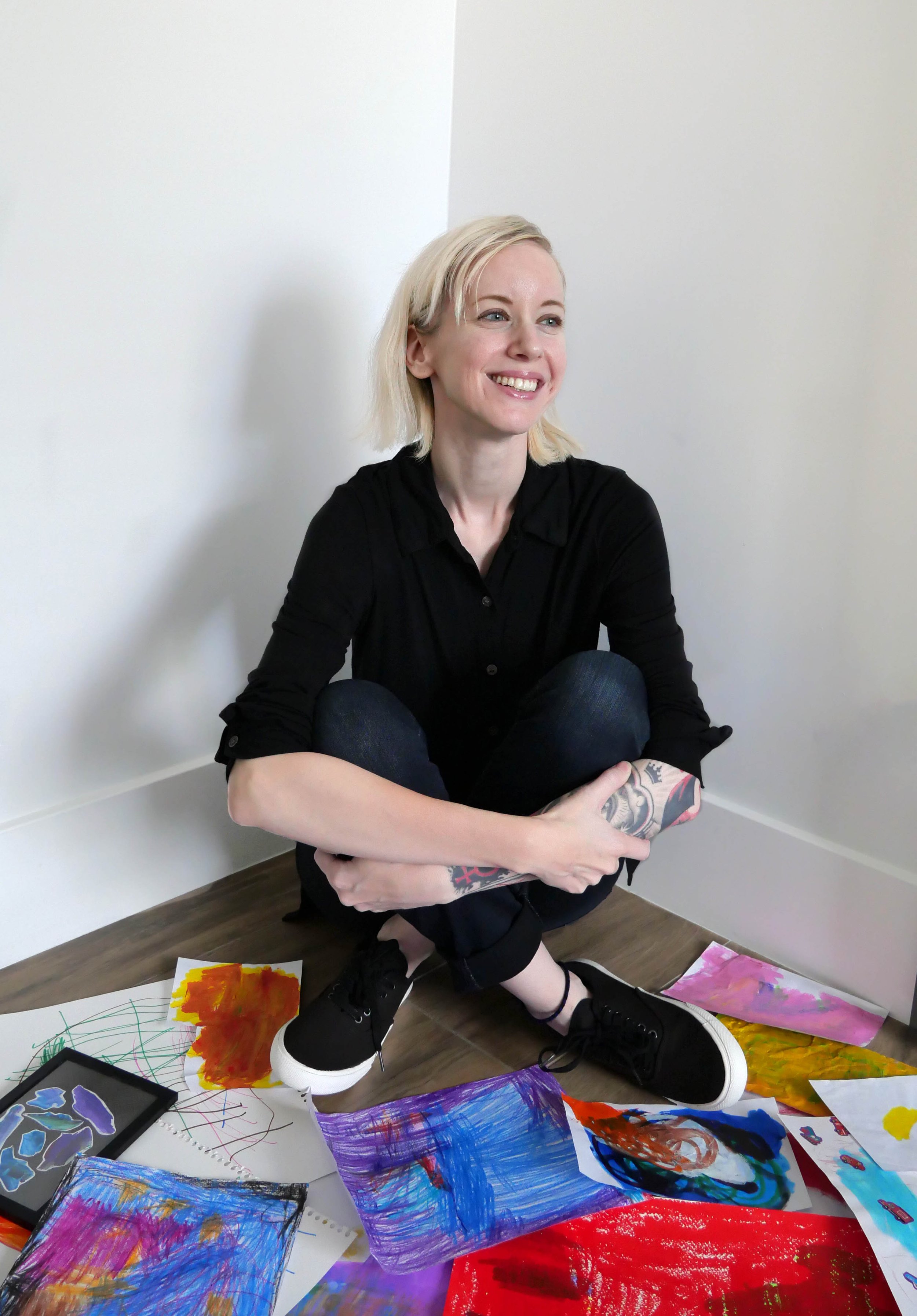 A woman with short blonde hair sitting on the floor, smiling, surrounded by colorful abstract paintings and drawings on paper.