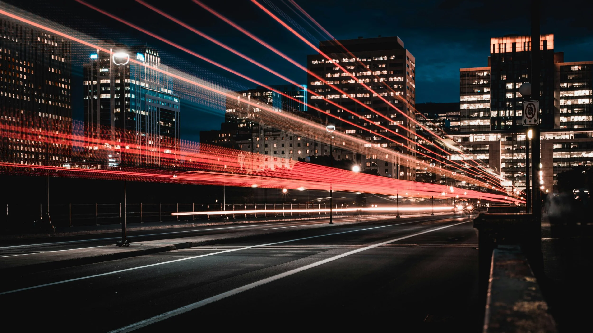 Nighttime cityscape with illuminated high-rise buildings and light trails from moving vehicles on the street.