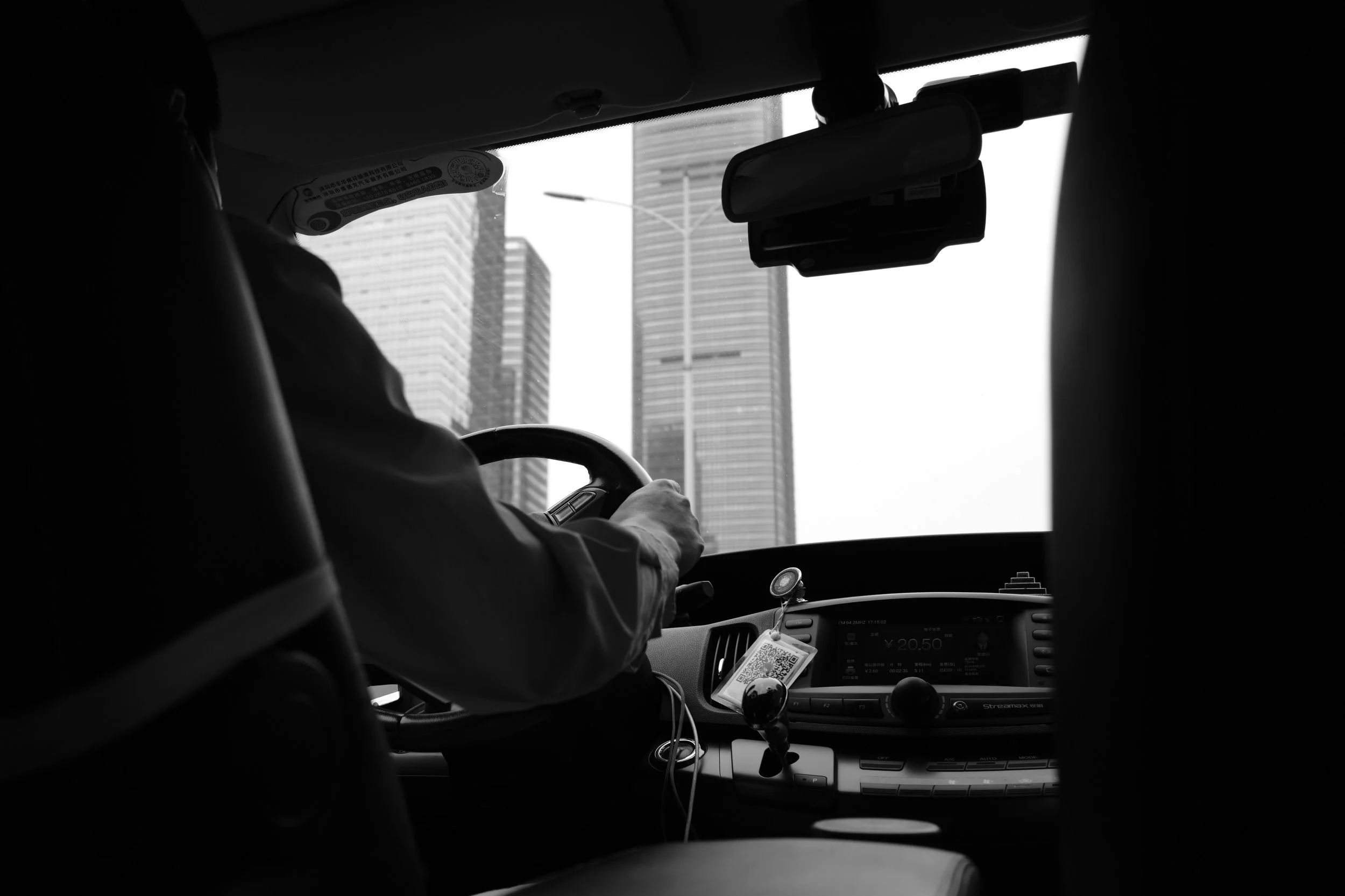 Inside a vehicle, the driver is visible with one hand on the steering wheel. The dashboard displays a radio and various controls. Outside the front windshield, tall city buildings are visible.
