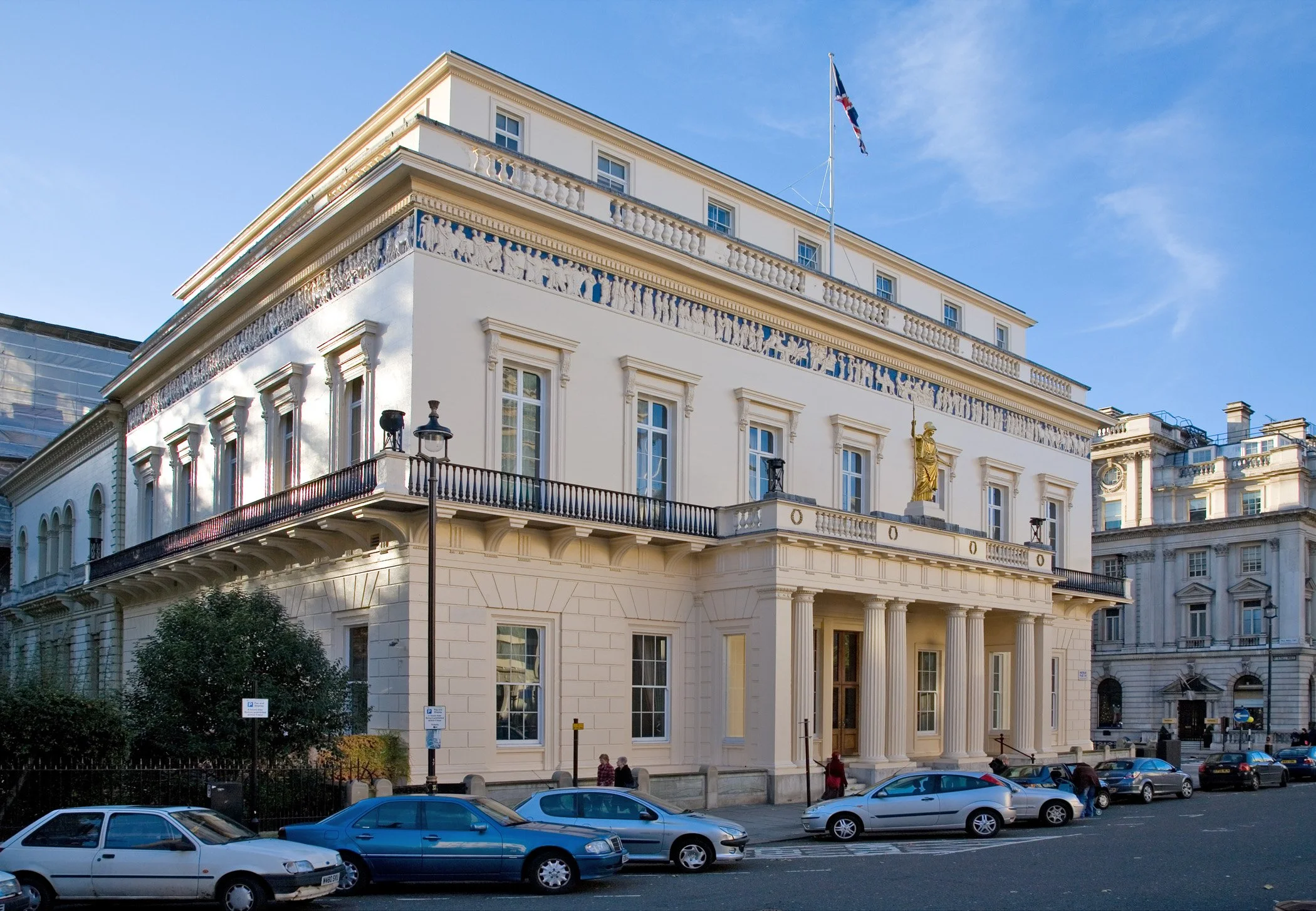 A white historic building with classical architectural features, including columns and decorative friezes, located on a city street with cars parked in front and a few pedestrians walking nearby. A flag is flying atop the building against a blue sky.
