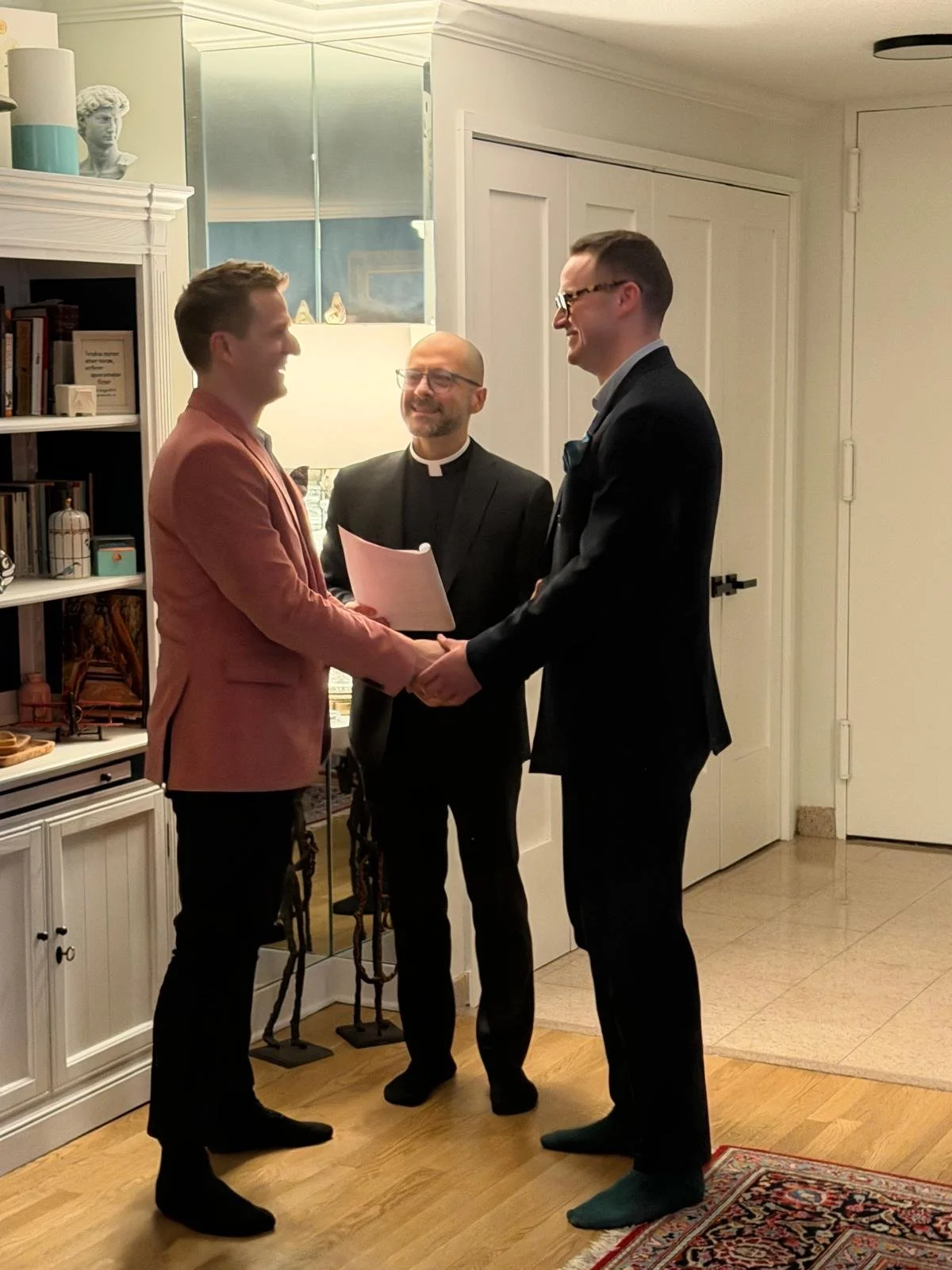 Two men in suits shaking hands during a wedding ceremony with a priest officiating, in a cozy indoor setting.