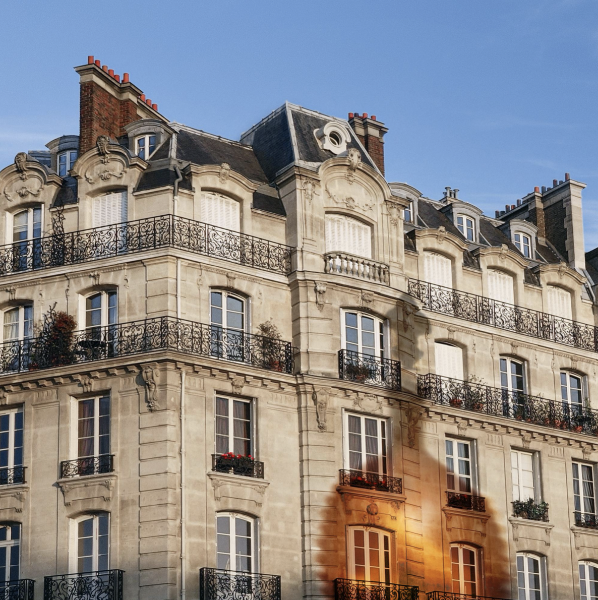 Immeuble haussmannien avec façades en pierre, balcons en fer forgé, fenêtres à volets et toits mansardés, sous un ciel clair.