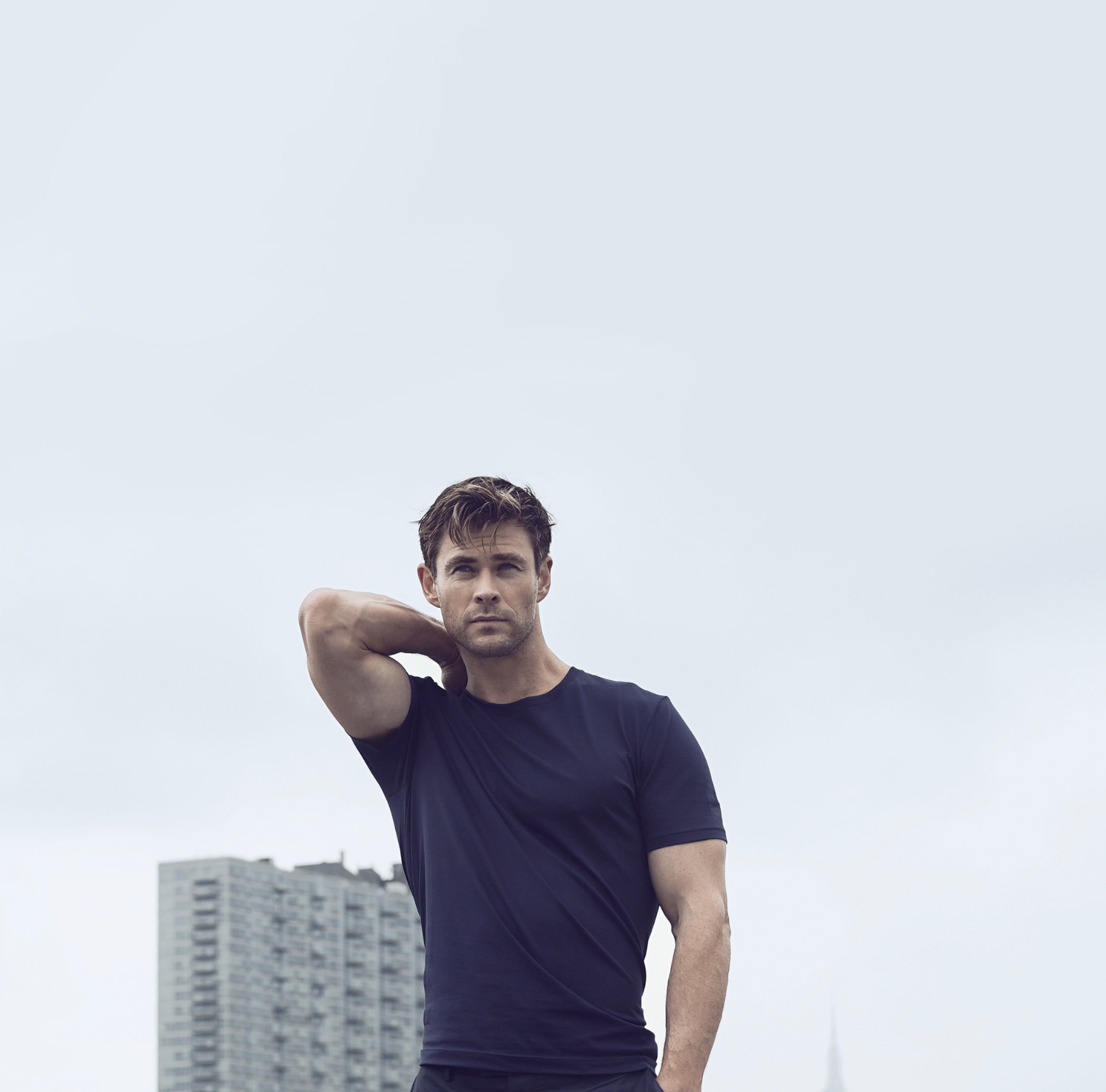 Un homme en t-shirt noir, debout devant un ciel nuageux avec un bâtiment en arrière-plan. Il regarde la caméra avec sérieux, une main derrière la tête.