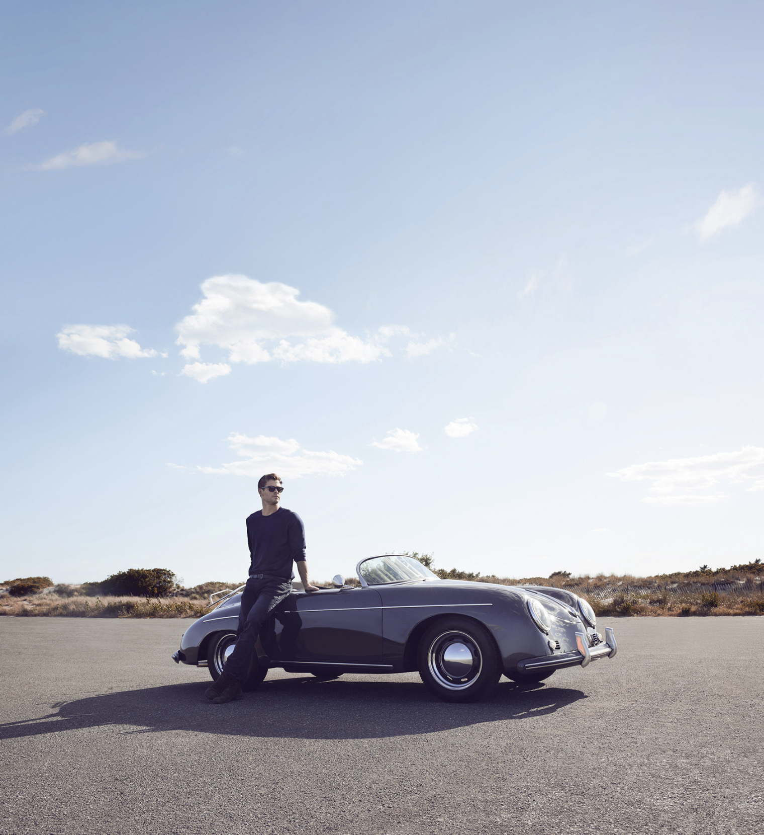 Un homme portant des lunettes de soleil, vêtu d'une chemise noire, est assis en biais sur une voiture de sport classique noire, dans un paysage ouvert avec un ciel bleu clair et des nuages sombres, sur une route ou un parking.