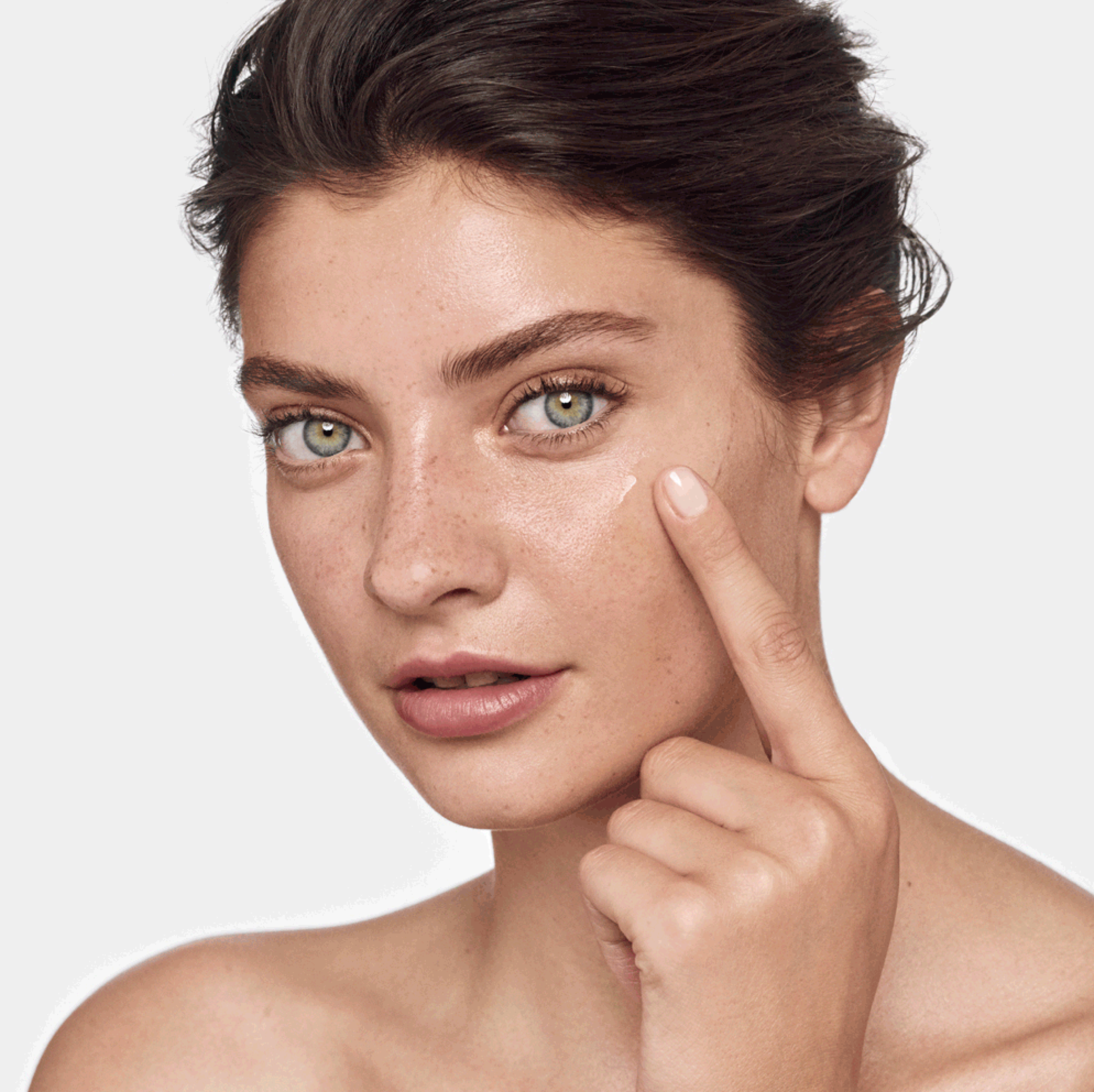 Une femme aux yeux vert clair, avec des cheveux courts et bruns, applique une crème sur son visage.