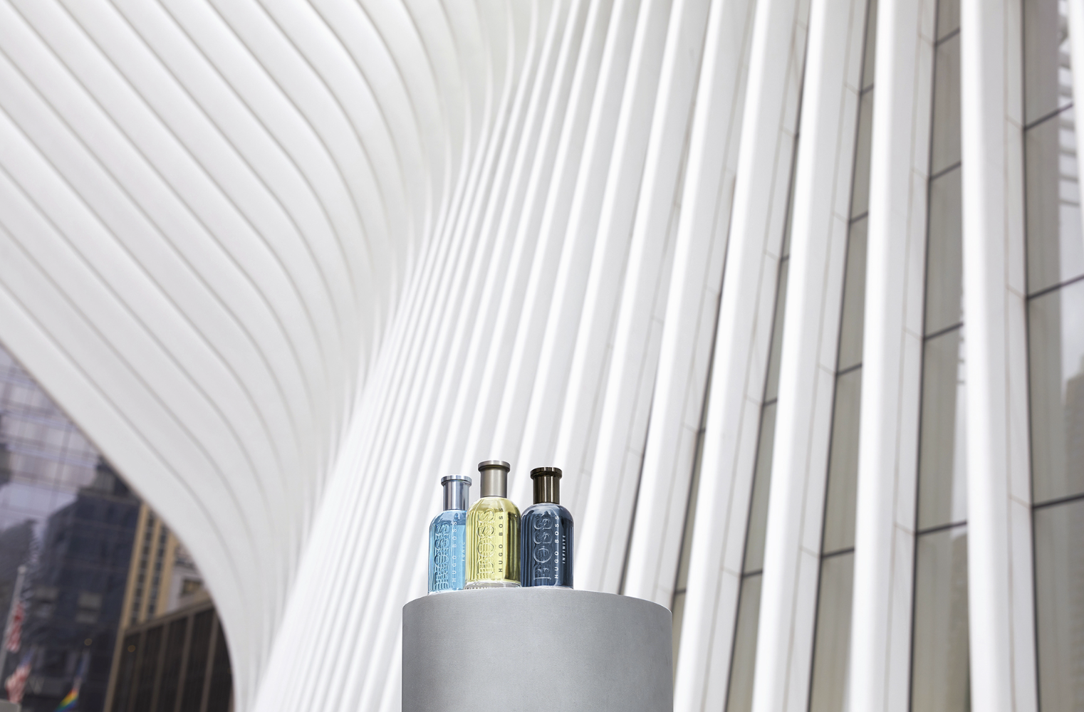 Trois bouteilles de parfum en différentes couleurs placées sur un piédestal dans un bâtiment moderniste avec des éléments architecturaux en blanc.