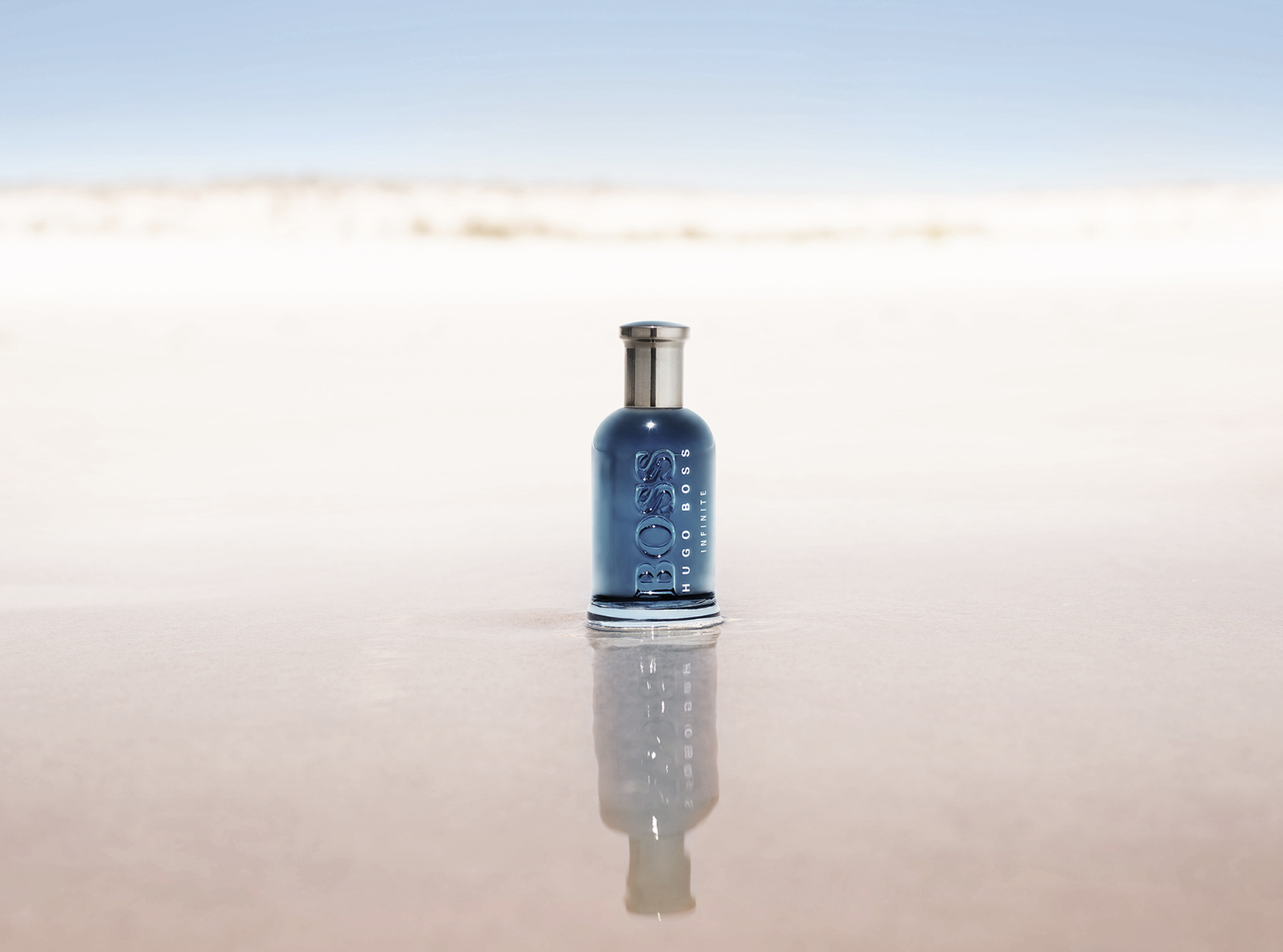 Flacon de parfum Hugo Boss Infinite en bleu sur une surface réfléchissante, plage en arrière-plan avec ciel clair.