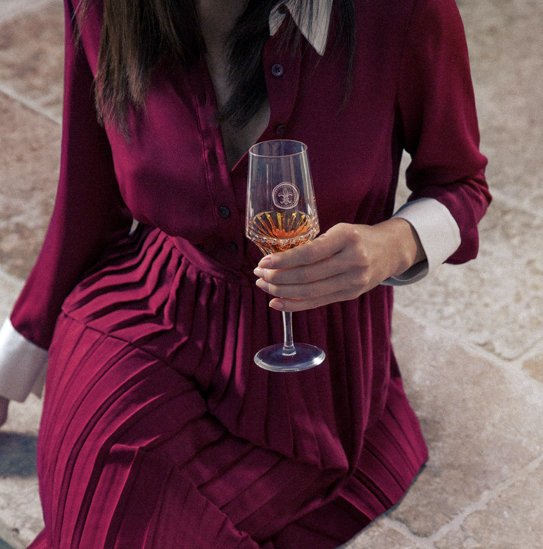 Une personne vêtue d'une robe rouge foncé avec une chemise blanche, tenant un verre à vin avec une boisson rosée, assise sur le sol en pierre.