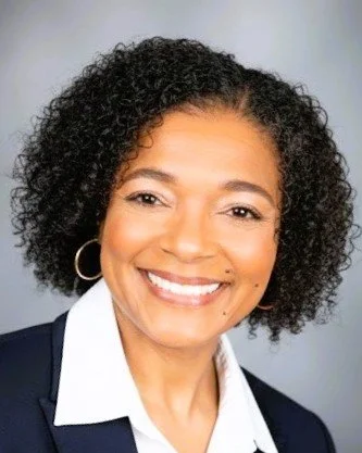 Smiling woman with curly black hair, wearing a white collared shirt and a dark blazer, against a neutral background.