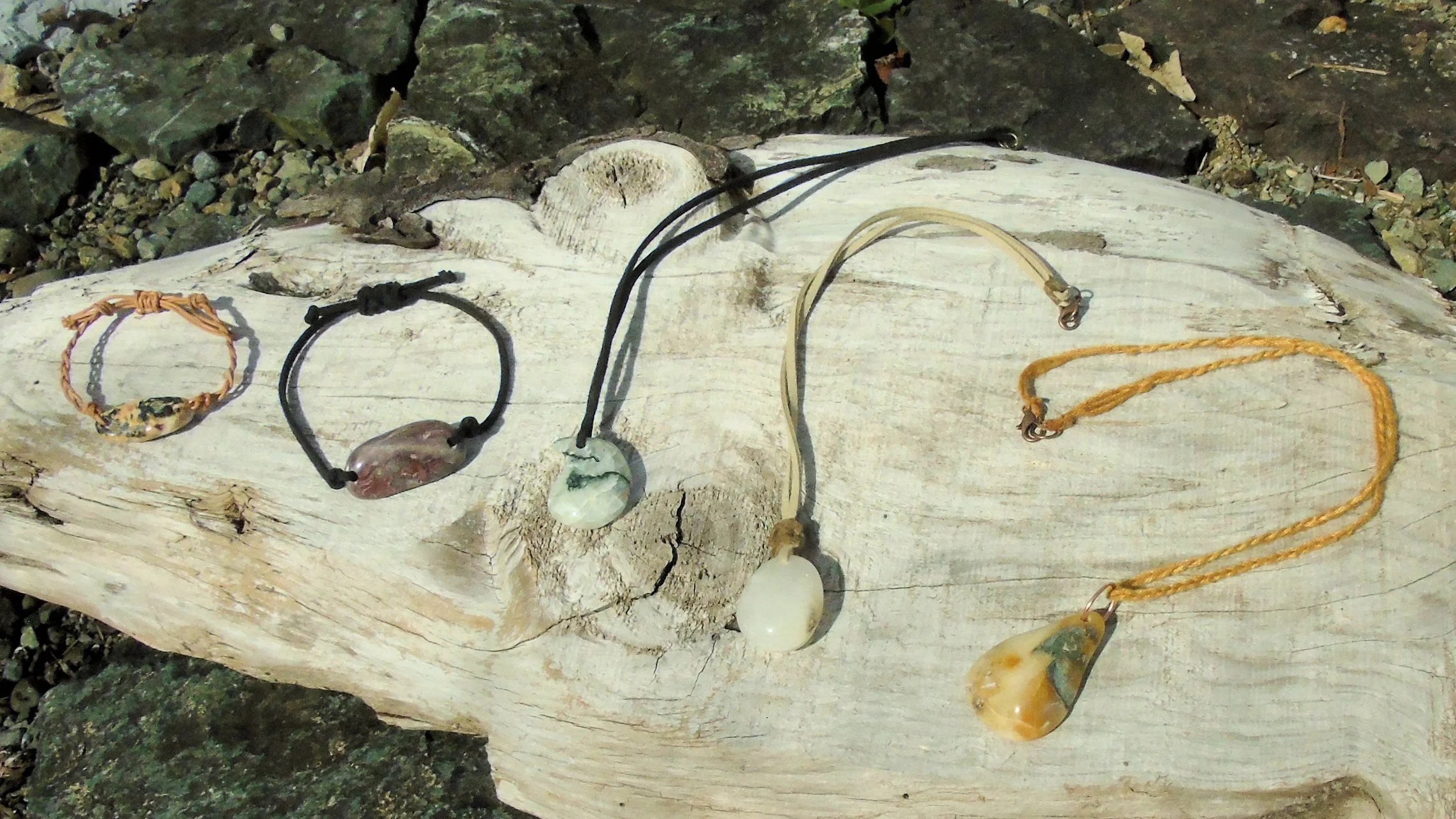 Various necklaces made from carved stones or shells draped on a weathered piece of driftwood, positioned outdoors on a rocky surface.