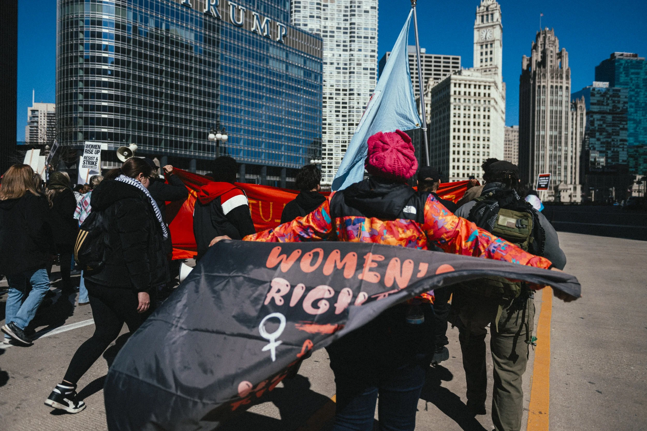 A group of protesters march in an urban area with tall buildings in the background, holding signs and banners advocating for women's rights.