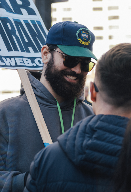 A man with a beard wearing sunglasses, a blue cap with an emblem, and a gray hoodie, smiling and talking to another person during a protest or rally, holding a sign that reads 'MAR' and 'WEB.COM'.
