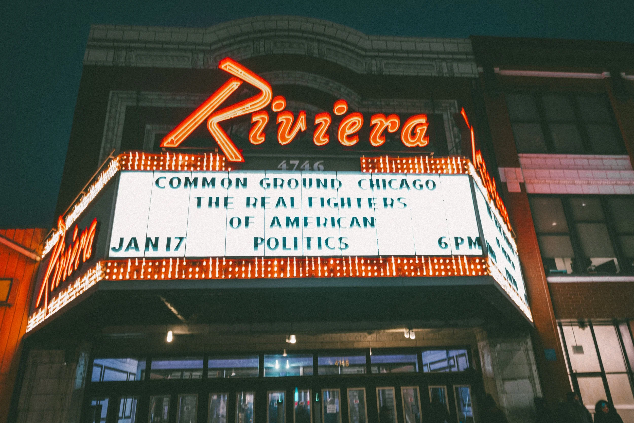 Night view of the Riviera Theatre marquee in Chicago, lit up in red neon, with a digital message board displaying event information.
