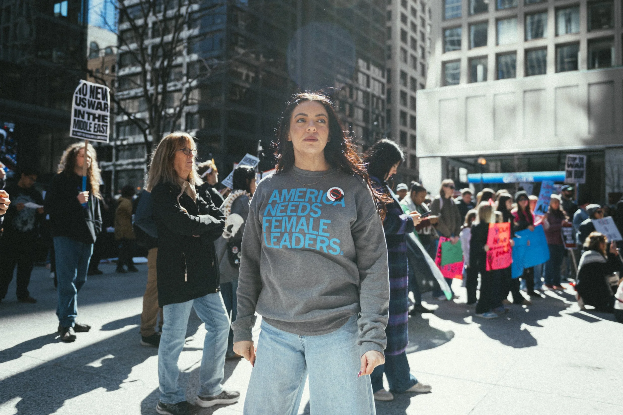 A woman walking in a crowd during a protest or rally in an urban cityscape. She wears a gray sweatshirt that reads 'America Needs Female Leaders' and light blue jeans, and has dark hair. People around her hold signs, including one that says 'No to New US War in the Middle East.'