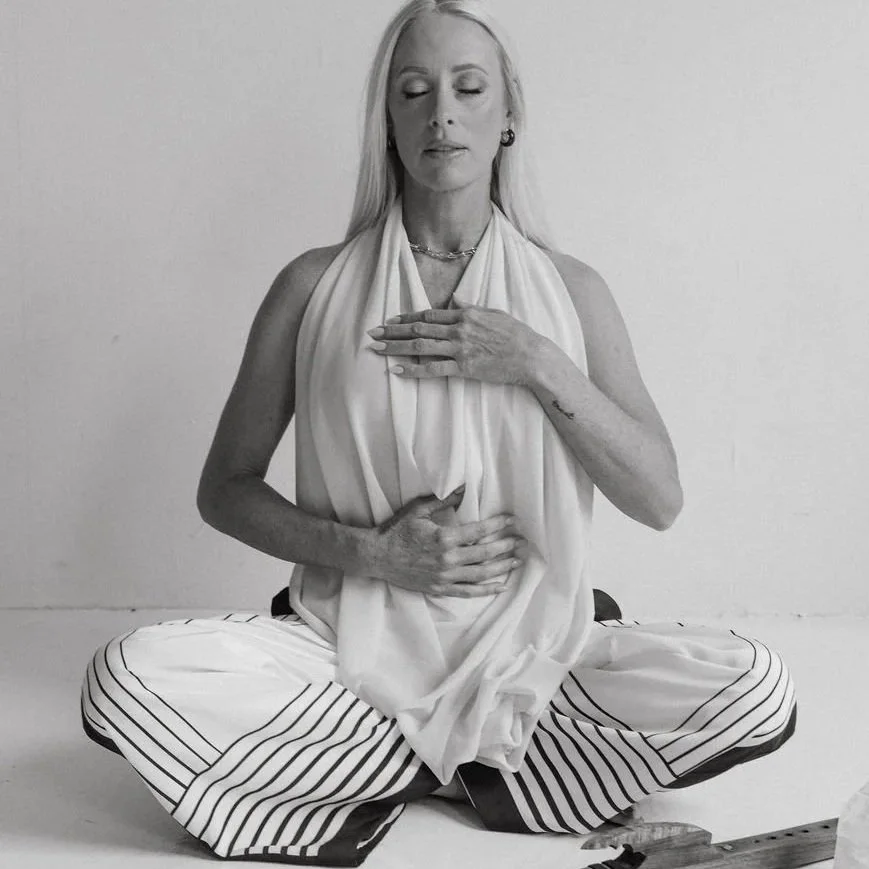 A woman sitting cross-legged with her eyes closed, wearing a white top and striped pants, placing one hand on her chest and one on her stomach, practicing meditation or mindfulness.