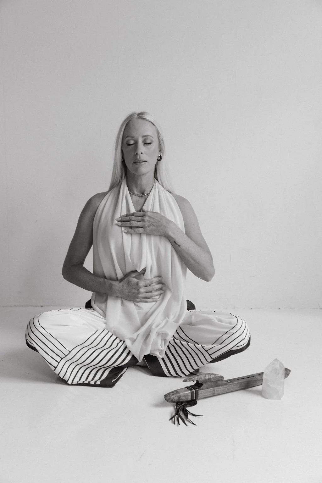 A woman with long blonde hair sits cross-legged on the floor with her eyes closed, wearing a sleeveless top and striped pants, holding her chest and stomach with her hands. In front of her, there is a crystal and a small wooden object with a tied leather strap.