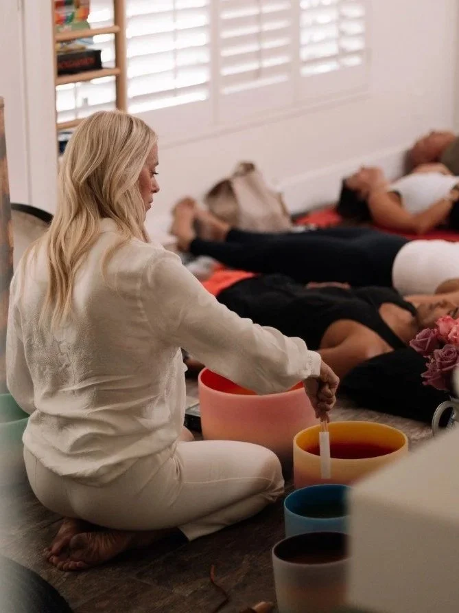 A woman with long blonde hair dressed in white is sitting on the floor with her legs crossed. She is holding a singing bowl and a stick, likely conducting a sound meditation. In the background, several people are lying down on mats with eyes closed, possibly participating in a meditation or relaxation session.