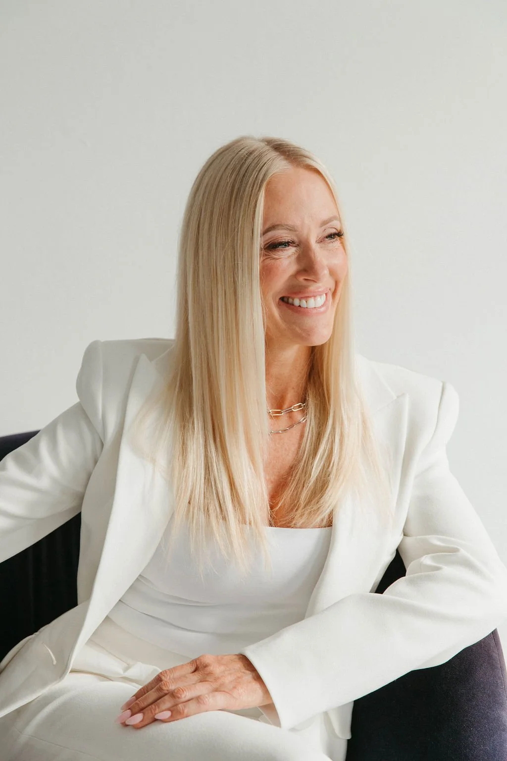 A woman with long blonde hair smiling, dressed in a white blazer and top, sitting on a dark chair against a plain white wall background.
