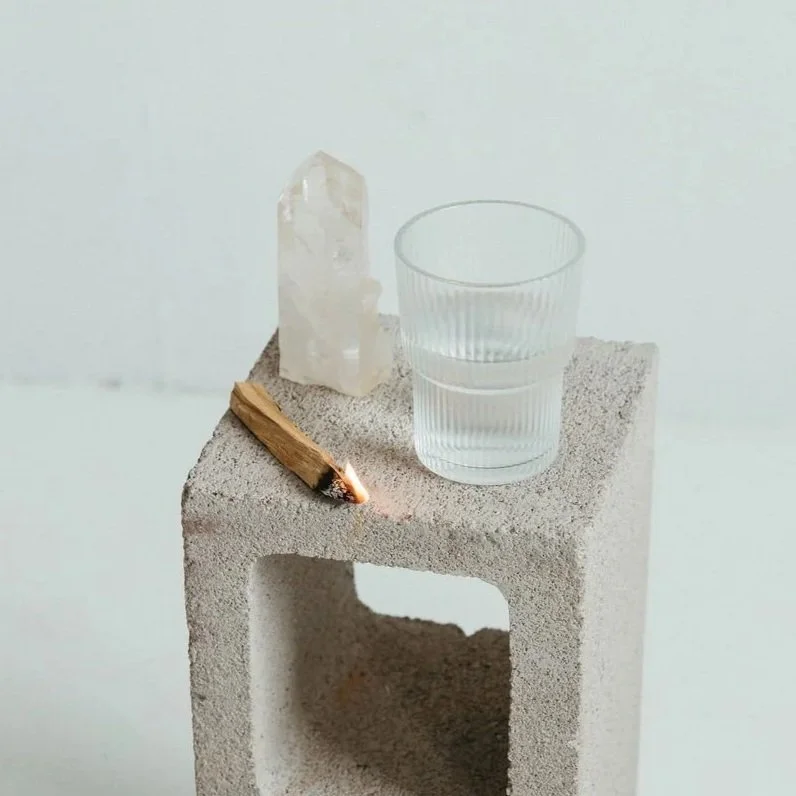 Clear glass of water, white crystal, and a lit wooden match on a concrete block.