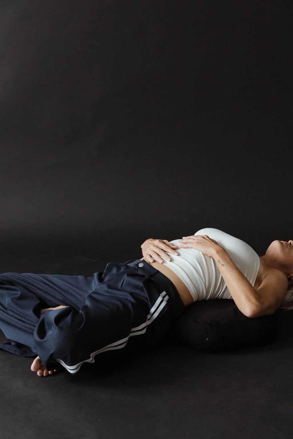 A person lying on their side on a black surface with a black background, wearing a white tank top and dark athletic pants.