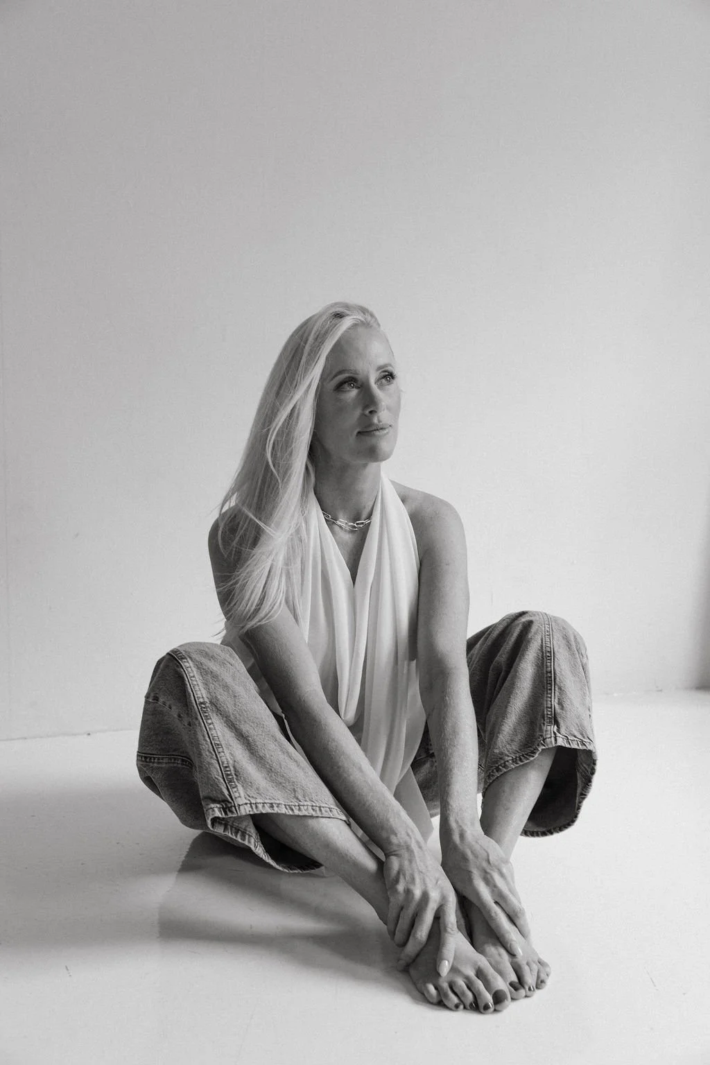 Black and white photo of a woman with long blonde hair, sitting cross-legged on the floor against a plain wall, looking away thoughtfully.
