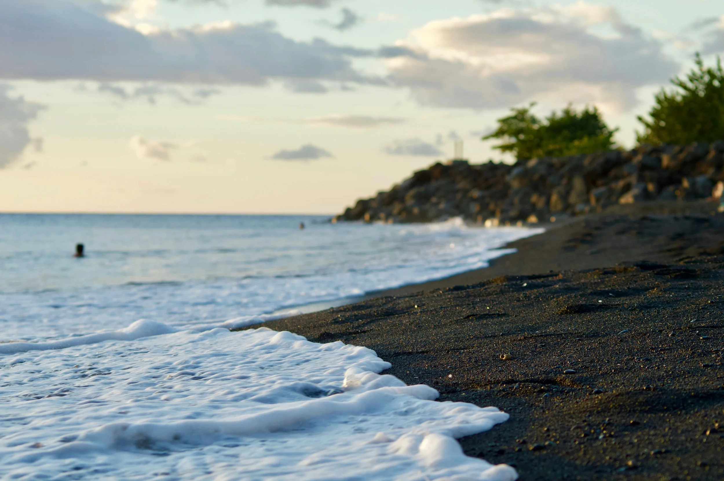 Sunset over a black sand beach with gentle waves, a rocky shoreline, and some trees in the distance.