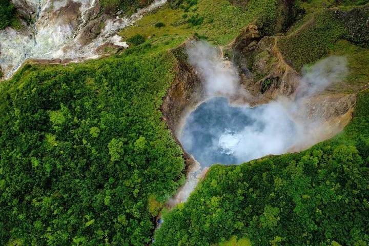 Aerial view of a geothermal hot spring crater with lush green forest surrounding it, emitting steam.