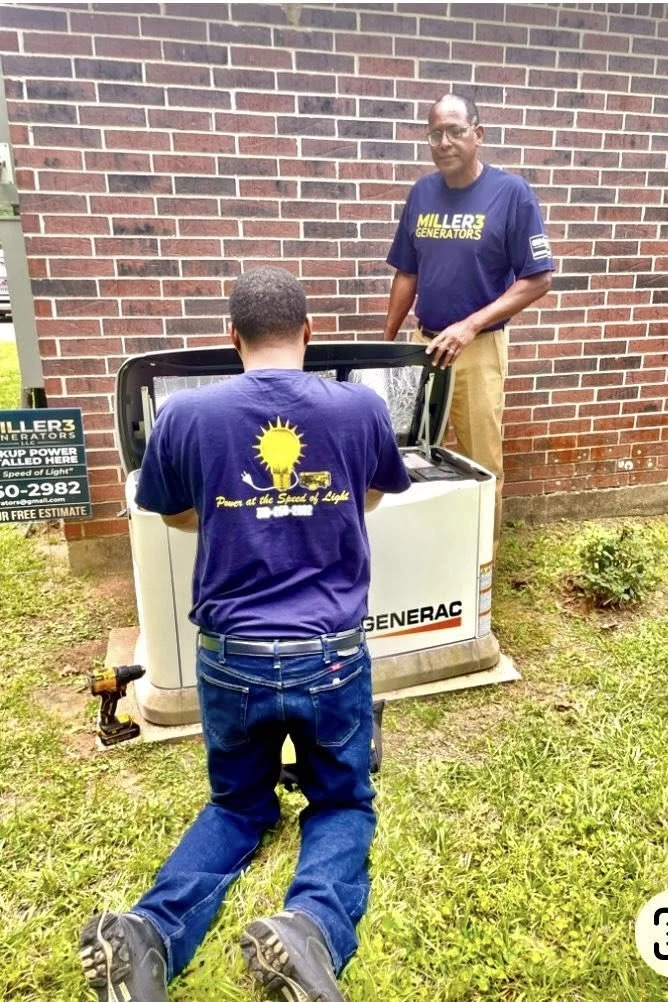 Two men installing or repairing a Generac generator outside a brick building, one standing and one kneeling on the grass.