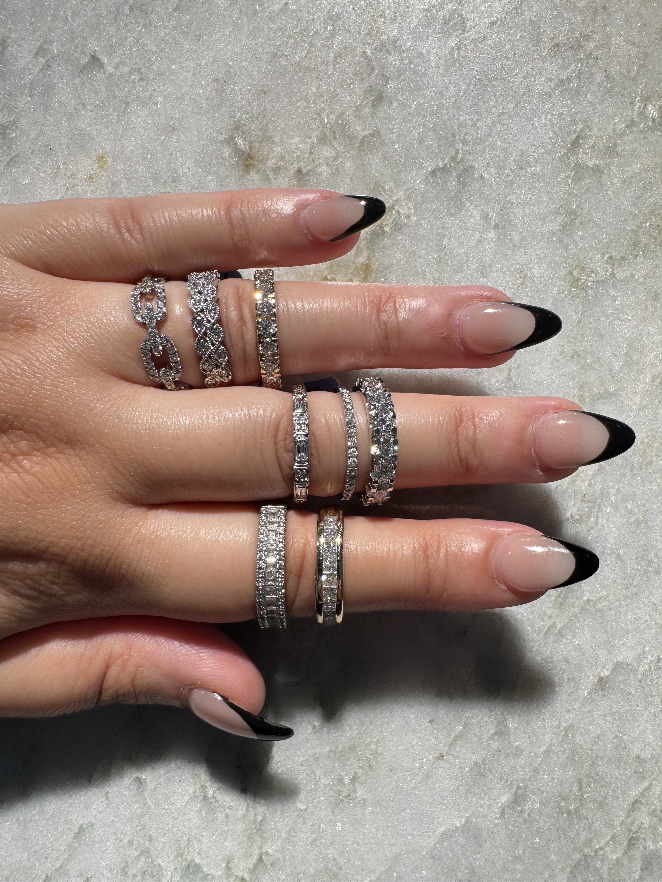 Close-up of a hand with multiple diamond rings, black French tip nails, resting on a white textured surface.