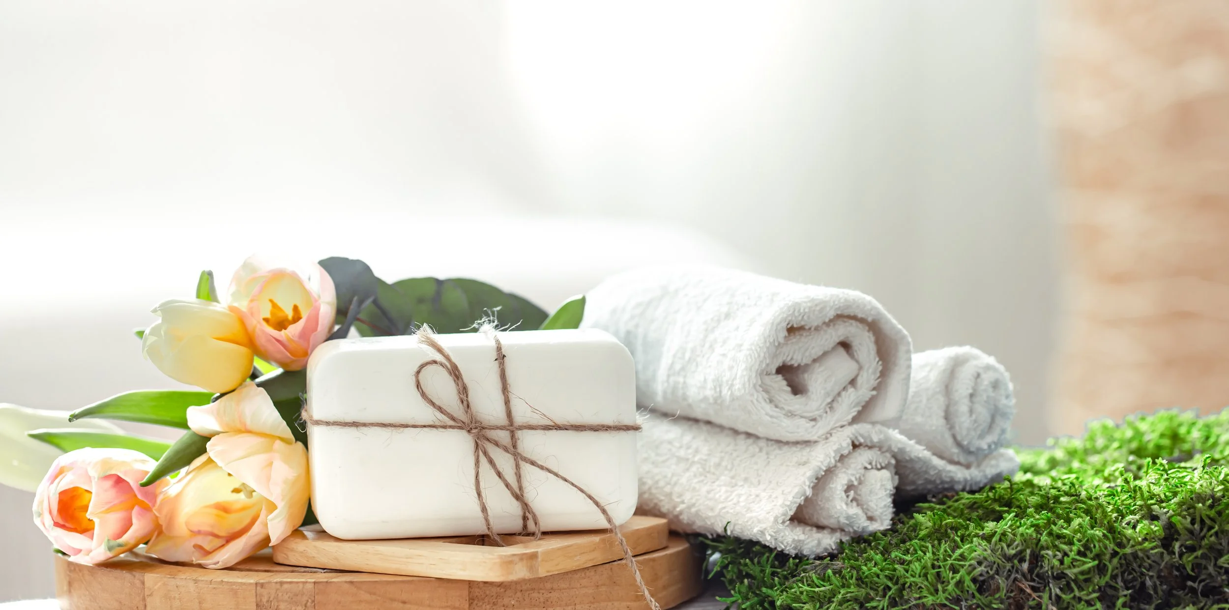 Décor de spa avec des serviettes blanches enroulées, des fleurs roses et jaunes, un savon blanc avec une ficelle en jute, et des plantes vertes en arrière-plan.