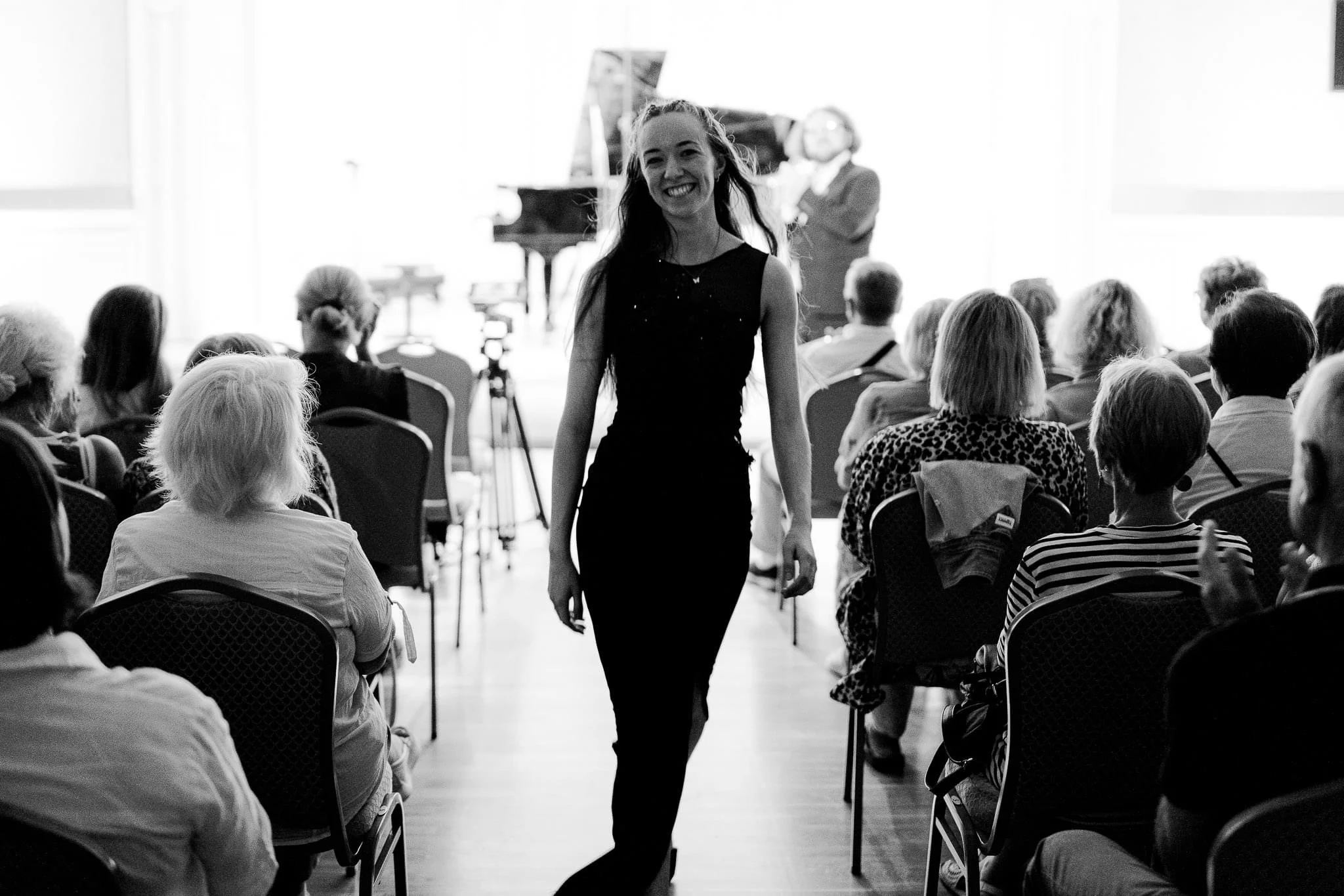 A woman in a sleeveless black dress walking down an aisle at a concert or event, with a piano and a person speaking in the background. Audience members are seated, facing the woman, with some looking towards her.