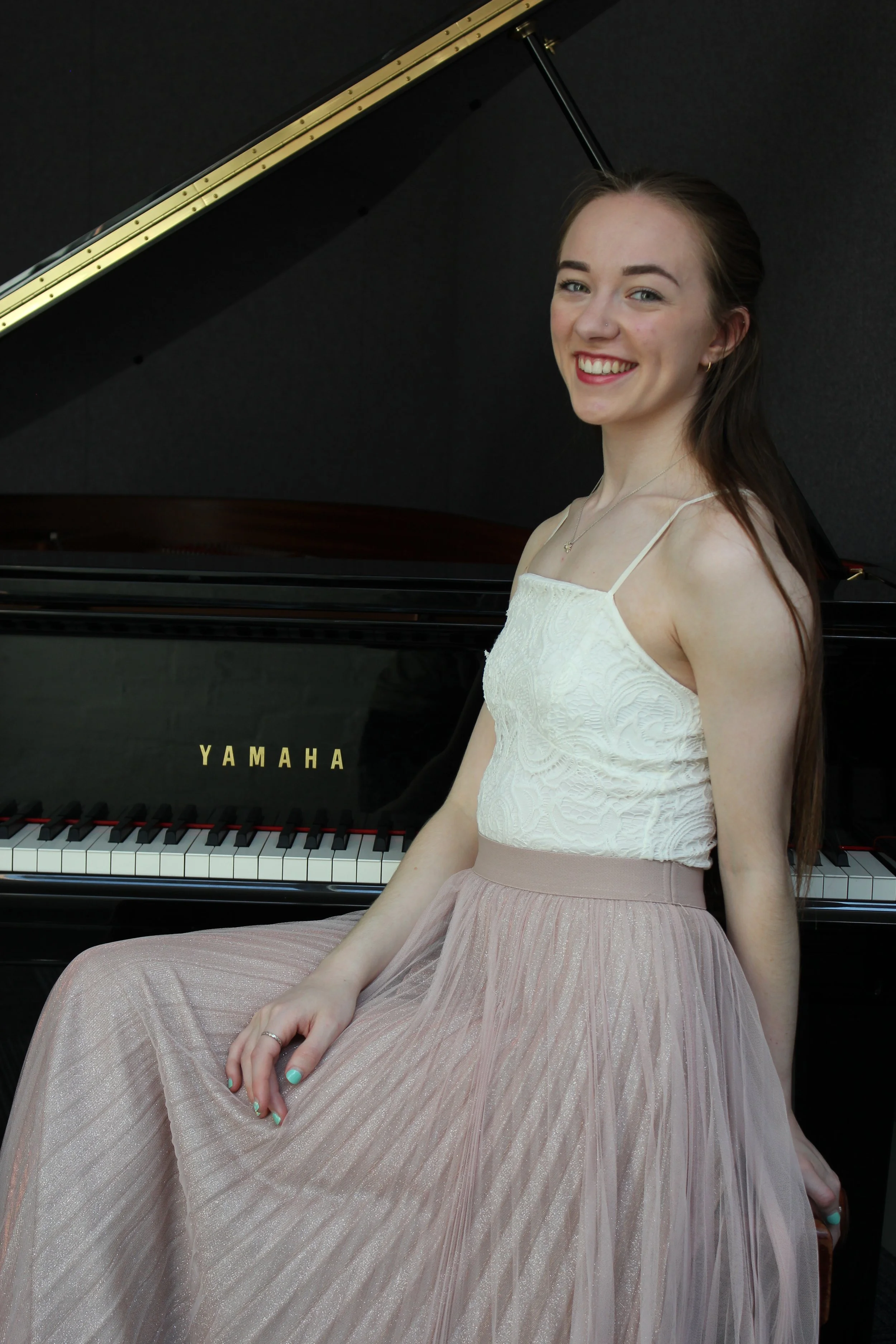 A young woman with long brown hair, smiling, sitting beside a black Yamaha grand piano, wearing a white spaghetti strap top and a pink tulle skirt.