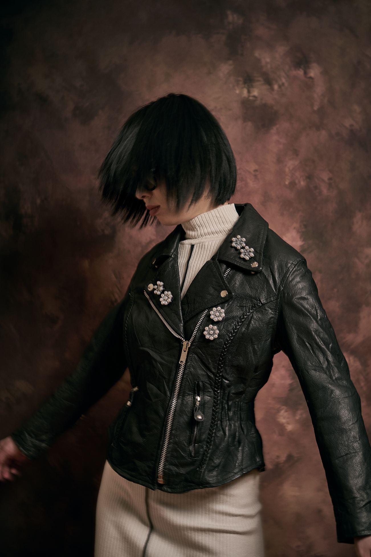 A person with short black hair covering their face, wearing a black leather jacket adorned with flower-shaped pearl embellishments, a white turtleneck, and beige pants, standing against a textured brown and pink background.