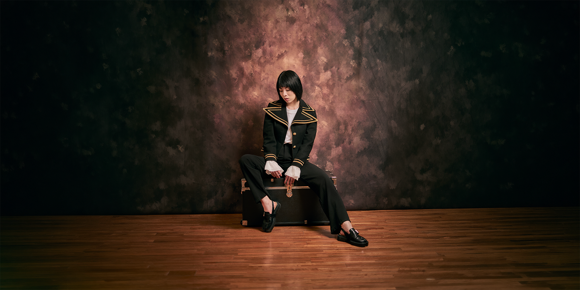 A woman in a black and gold jacket and black pants sits on a vintage trunk in a room with a dark, textured wall and hardwood floor.