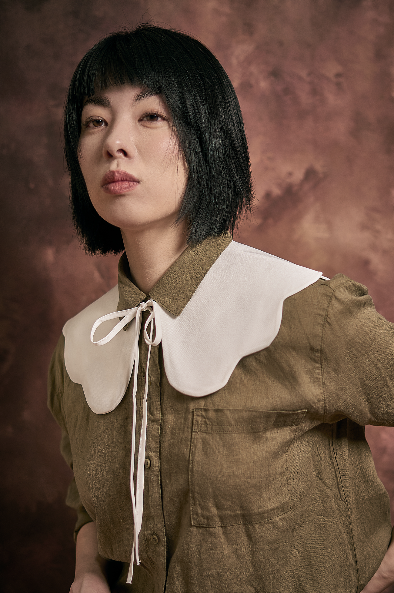 A woman with short black hair and a serious expression wears a brown shirt with a large white collar tied with a bow, standing against a brown textured background.