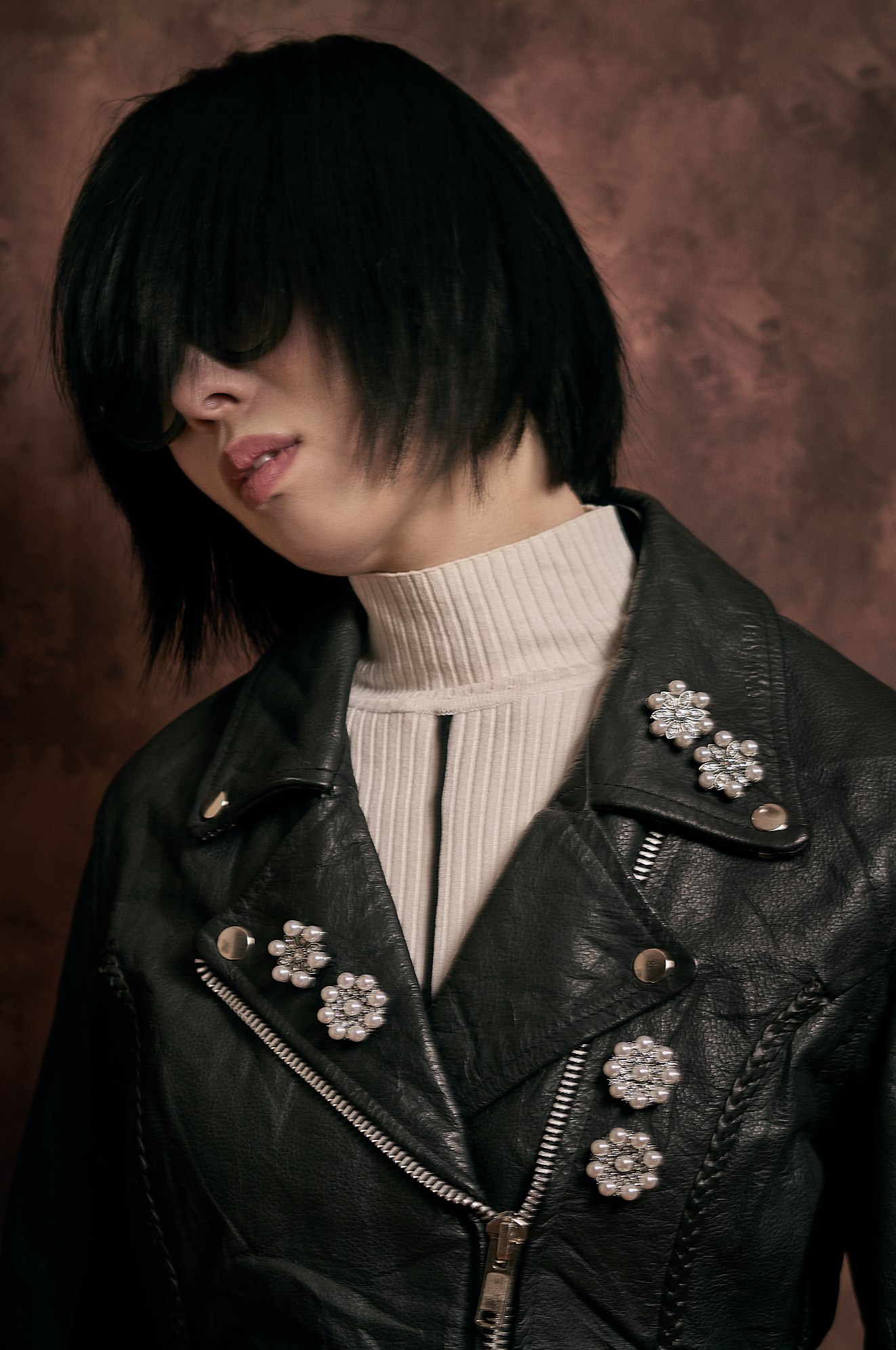 Woman with black hair, wearing sunglasses, a black leather jacket decorated with pearl and rhinestone floral embellishments, and a beige turtleneck shirt, against a brown background.