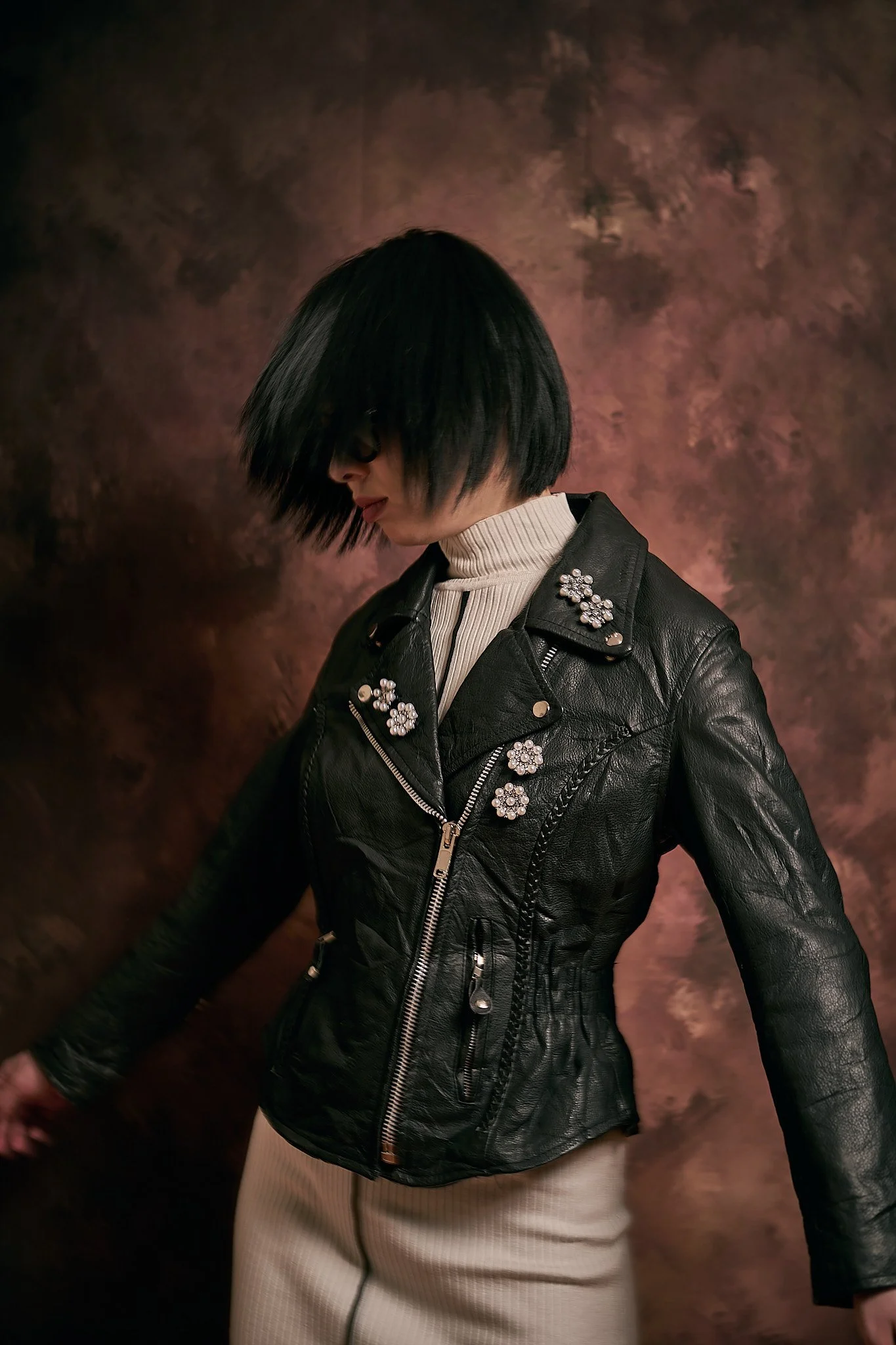 A woman with black, shoulder-length hair and wearing glasses is looking down. She is dressed in a black leather jacket with floral embellishments on the shoulders and a white ribbed turtleneck sweater underneath. The background is a textured brown backdrop.