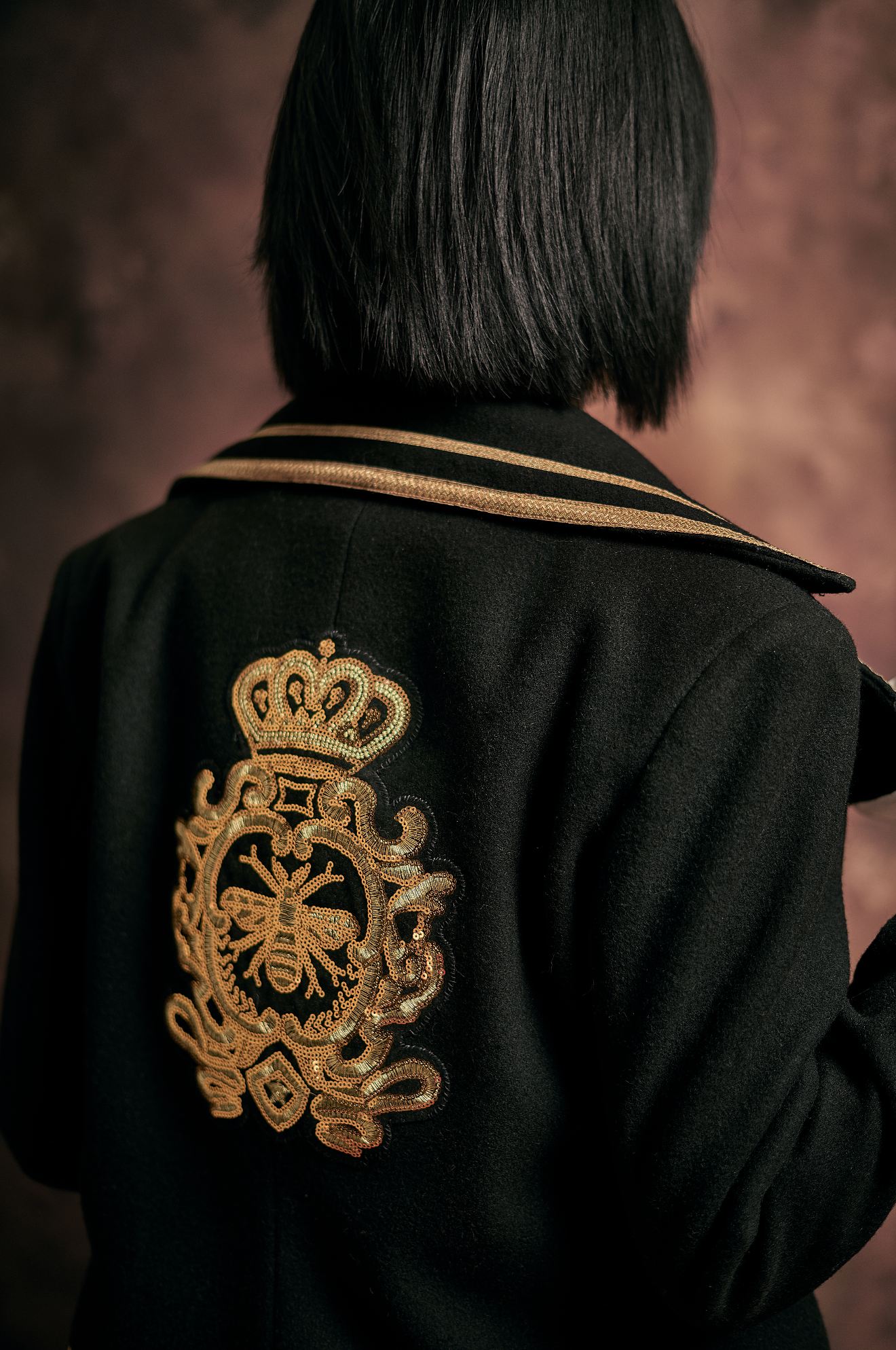 Back of a person with short black hair wearing a black uniform jacket with a gold embroidered crest on the sleeve, against a blurred pinkish background.