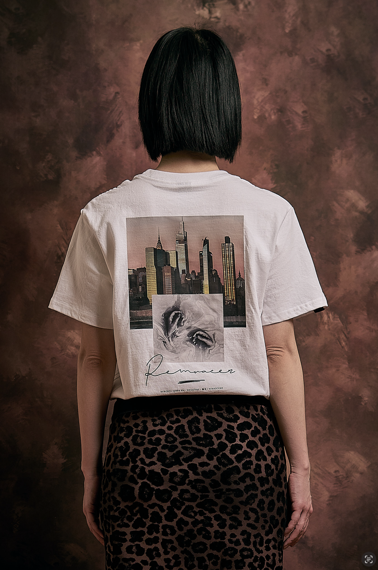 Back of a person with black hair wearing a white T-shirt featuring a city skyline and a drawing of a face, paired with leopard print pants, standing against a brown textured background.