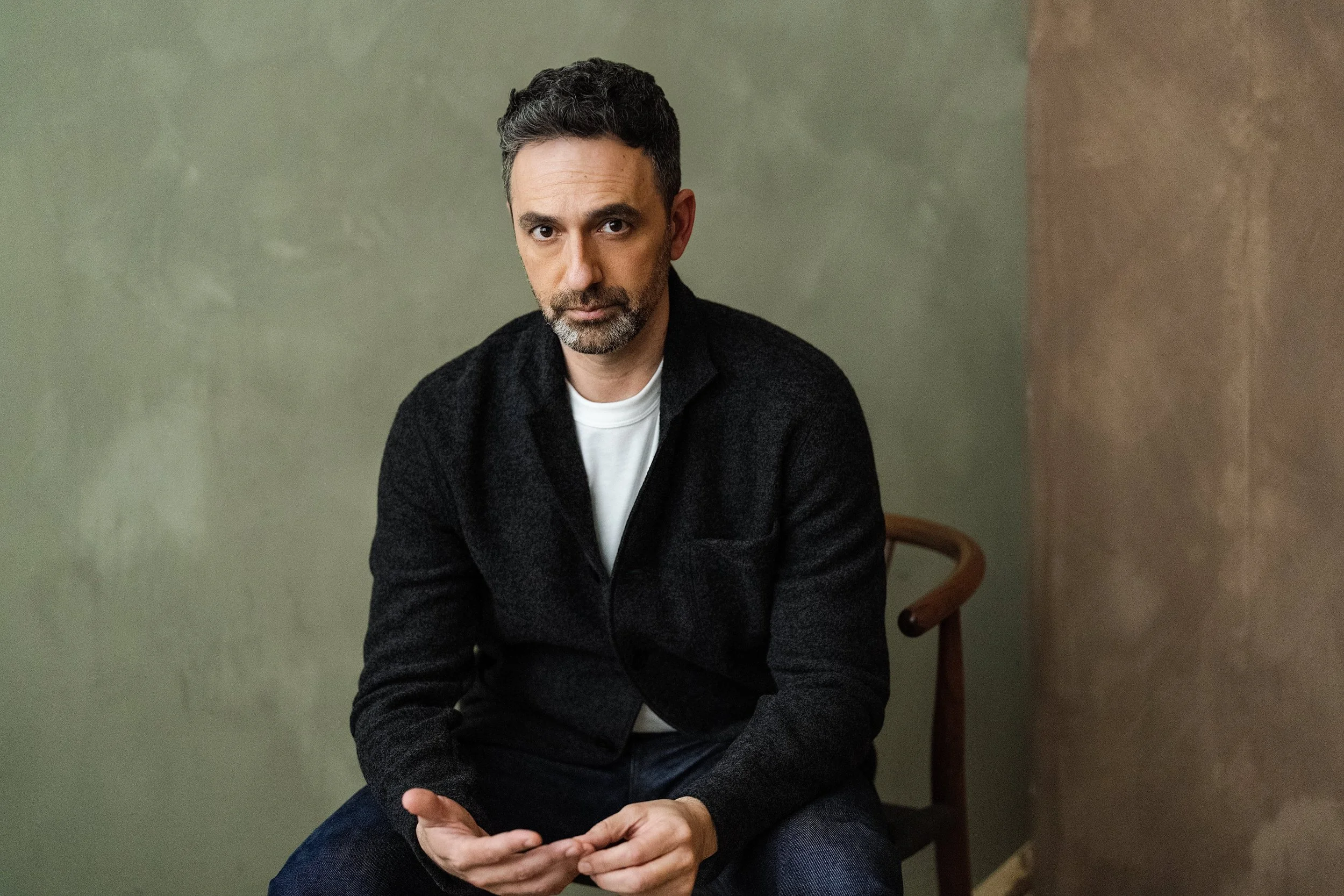 A man with dark hair and a beard sitting on a wooden chair against a muted green and brown wall, wearing a black jacket and white shirt.